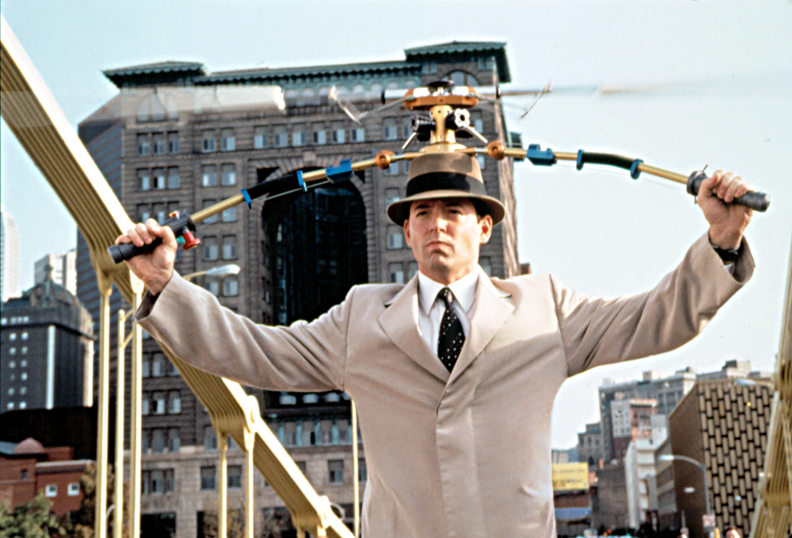 Matthew in a trench coat and hat with a copter gadget near a bridge and cityscape