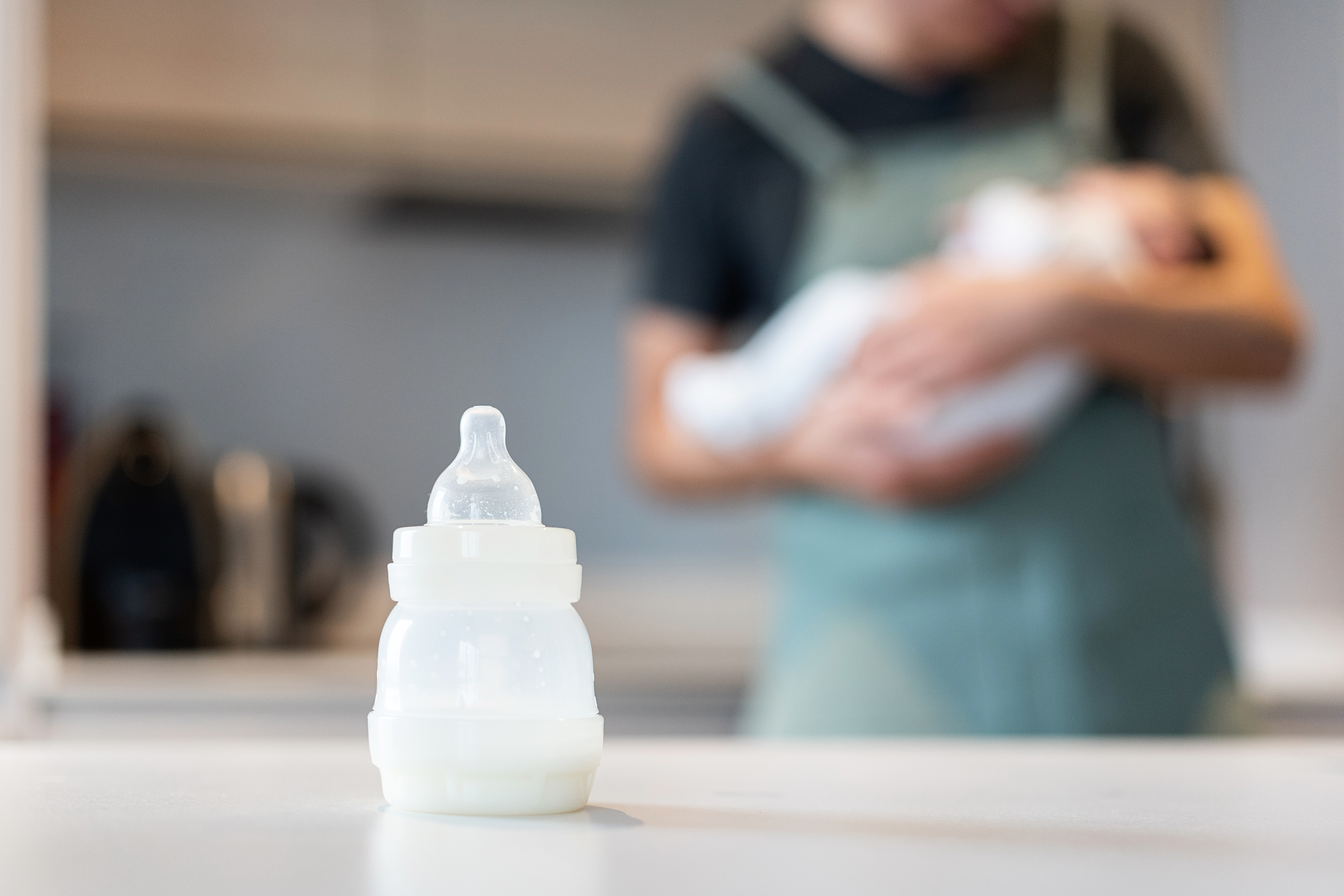 A baby bottle filled with milk is on a table. In the background, an unfocused person holds a baby