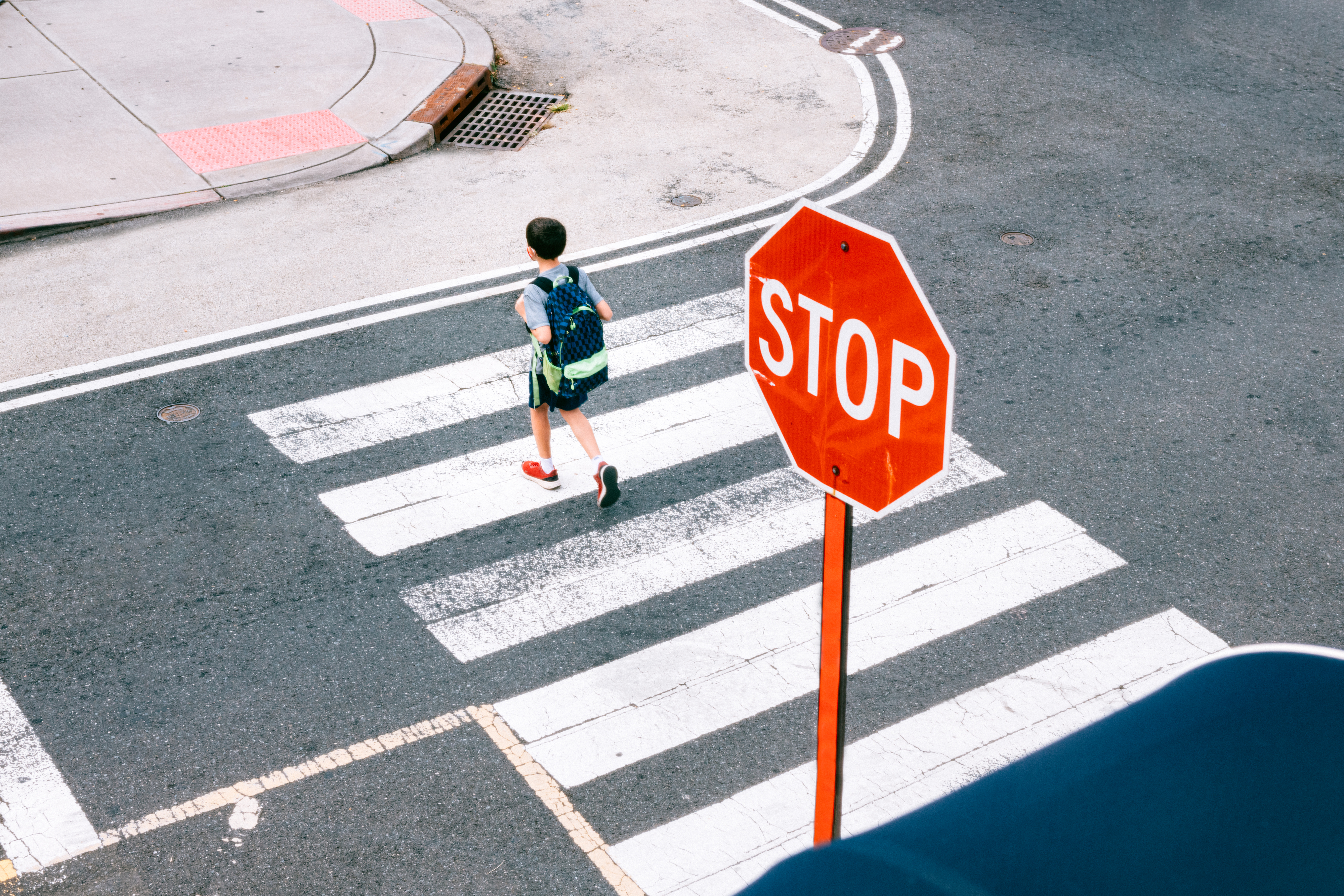 Child crossing street at crosswalk with a "STOP" sign nearby, wearing a backpack and sneakers. Urban street setting