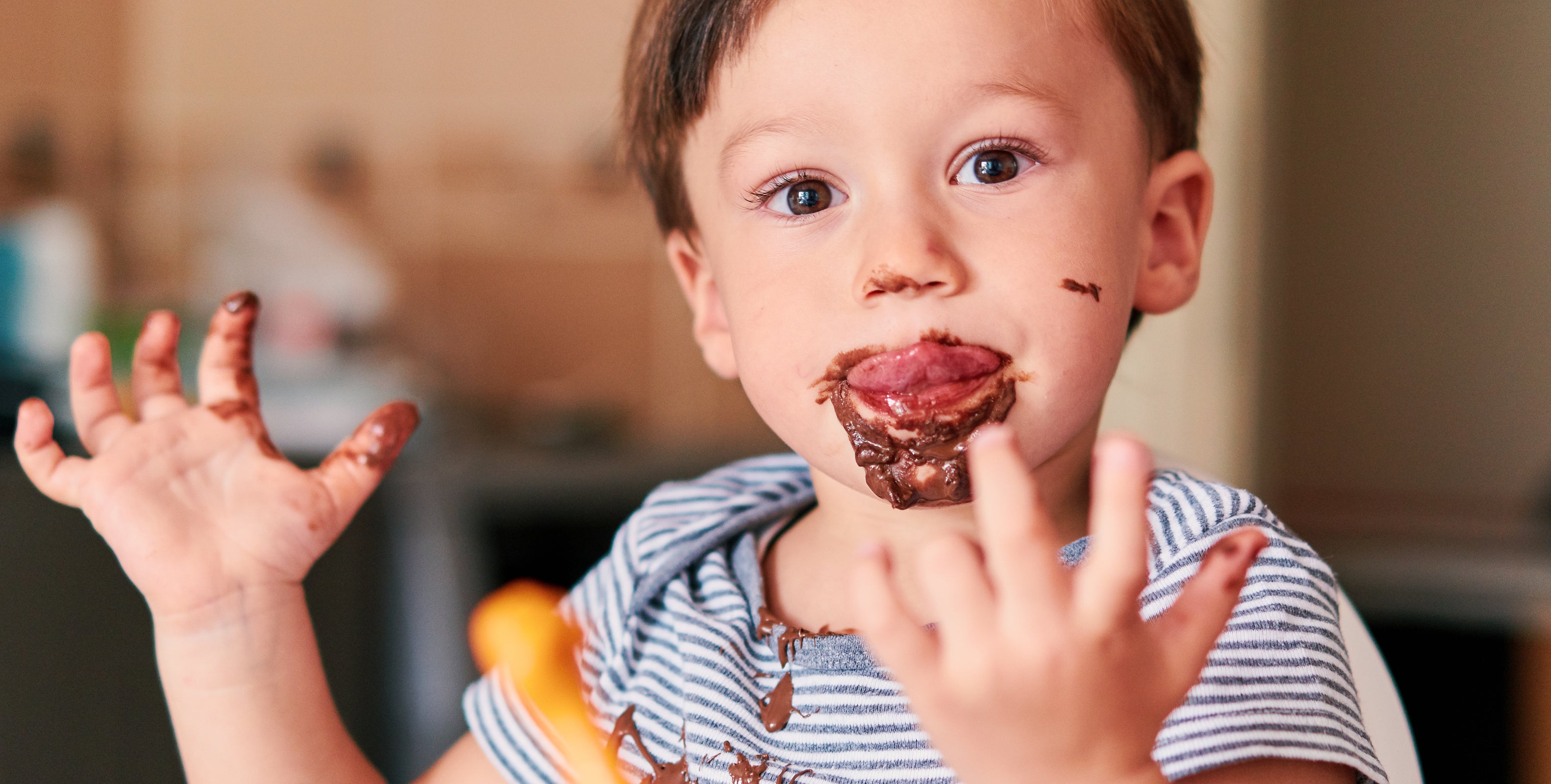 A child with short hair is sitting at a table with chocolate on their face and hands, looking curious and playful