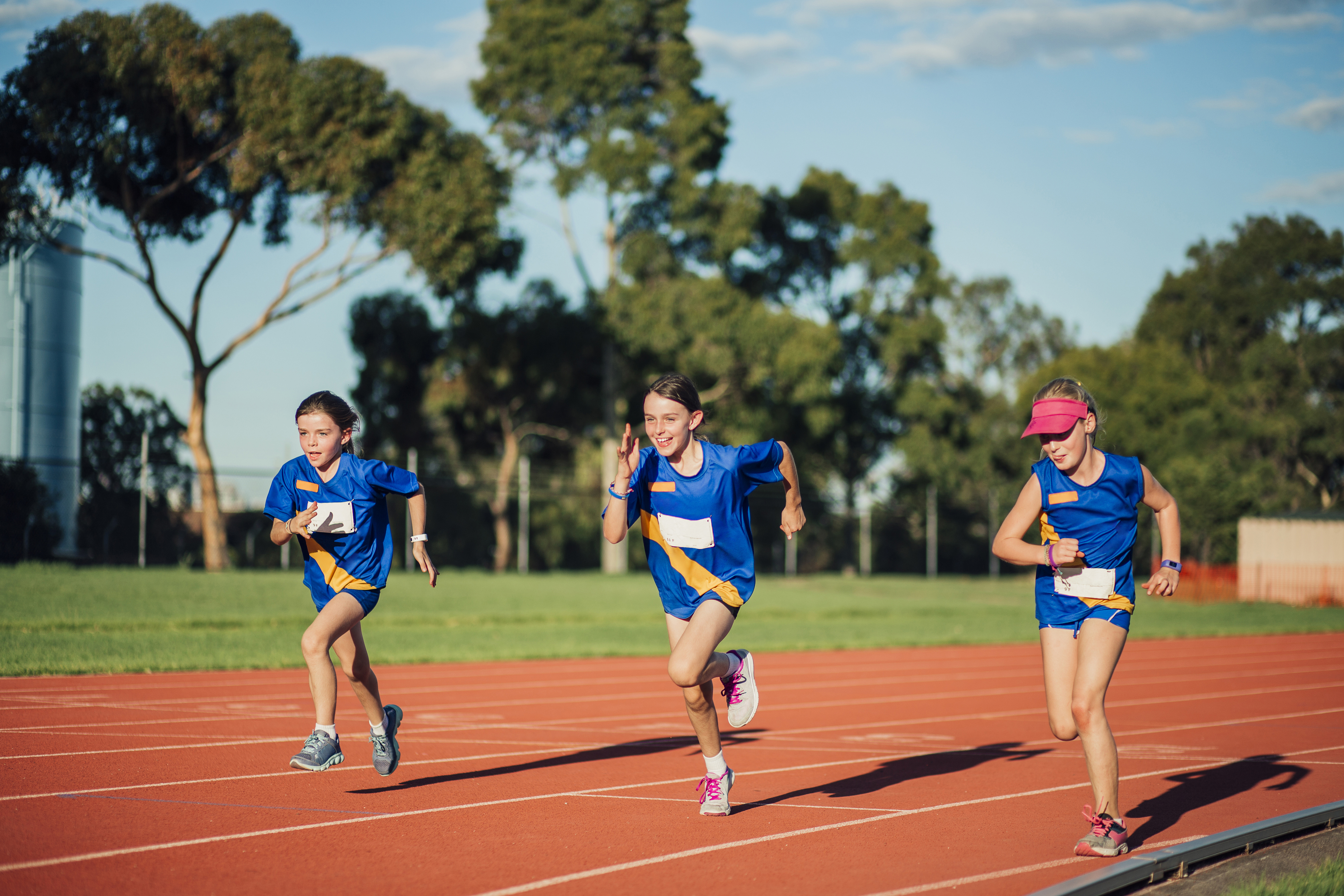 Three children sprint on an outdoor track, wearing blue athletic gear and race bibs. Trees and grass are visible in the background