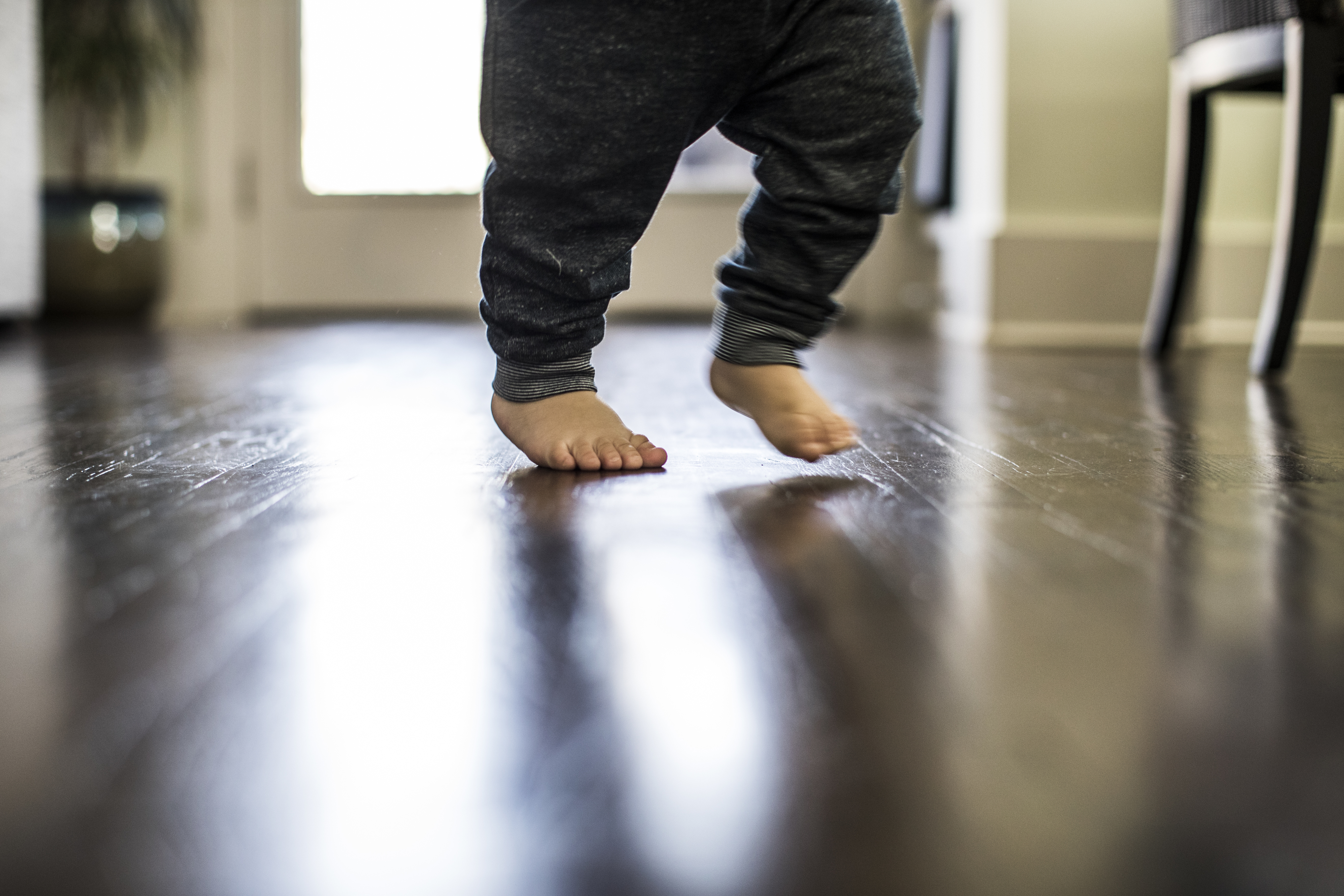 A toddler's bare feet take a step on a shiny wooden floor indoors