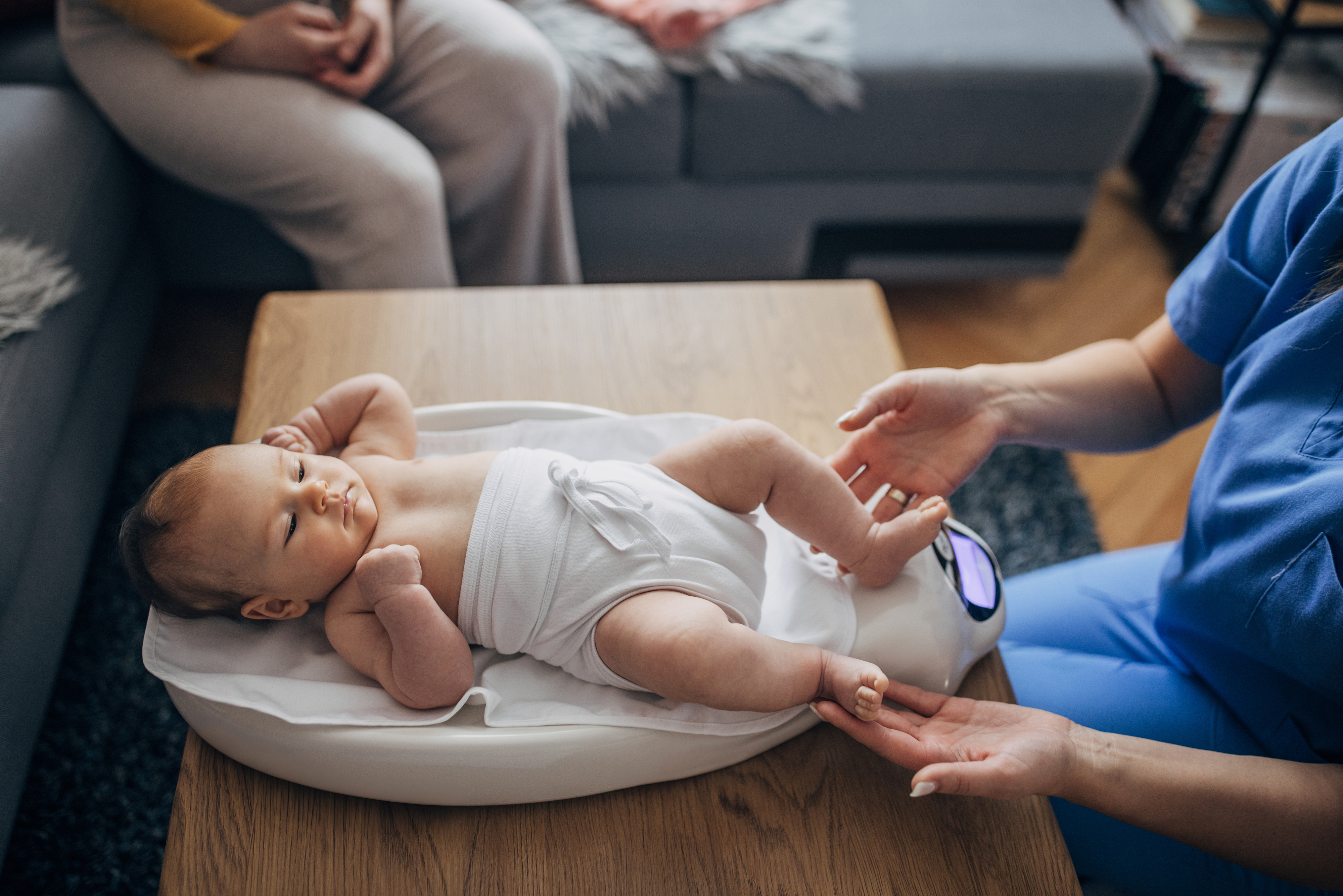 A nurse in scrubs checks a baby wrapped in a towel on a weighing scale, with a person sitting in the background