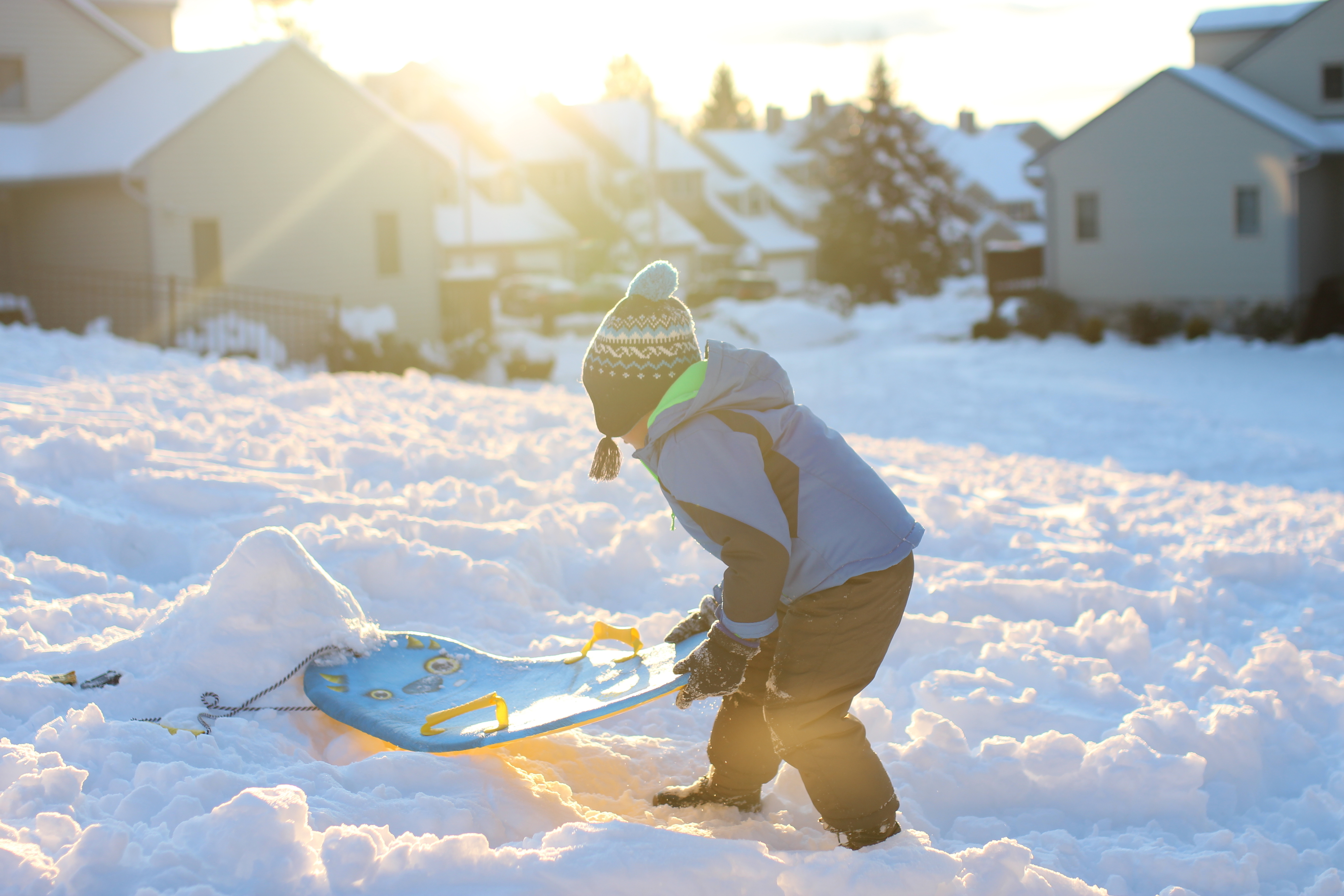Child wearing winter clothing plays with a sled in a snowy neighborhood. Bright sunlight is in the background