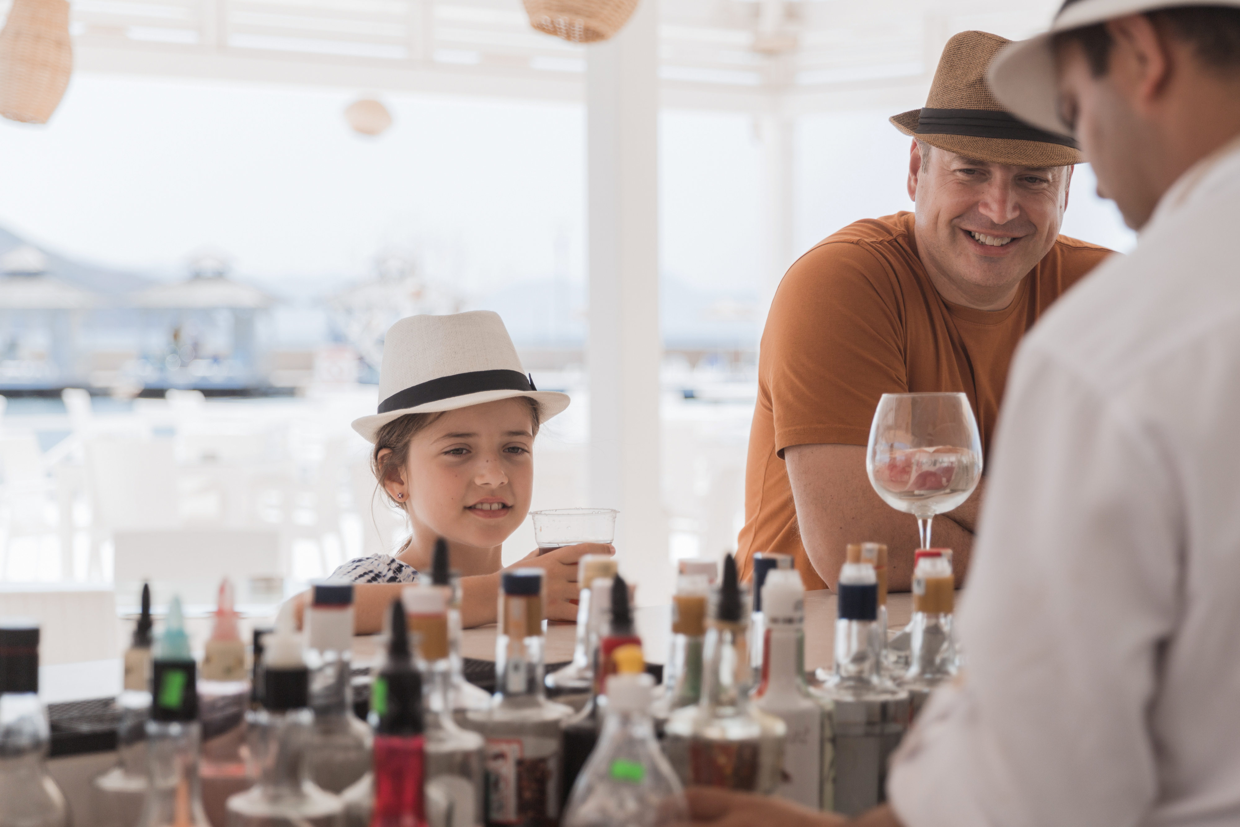 A child and an adult in hats enjoy drinks at an outdoor bar, served by a bartender