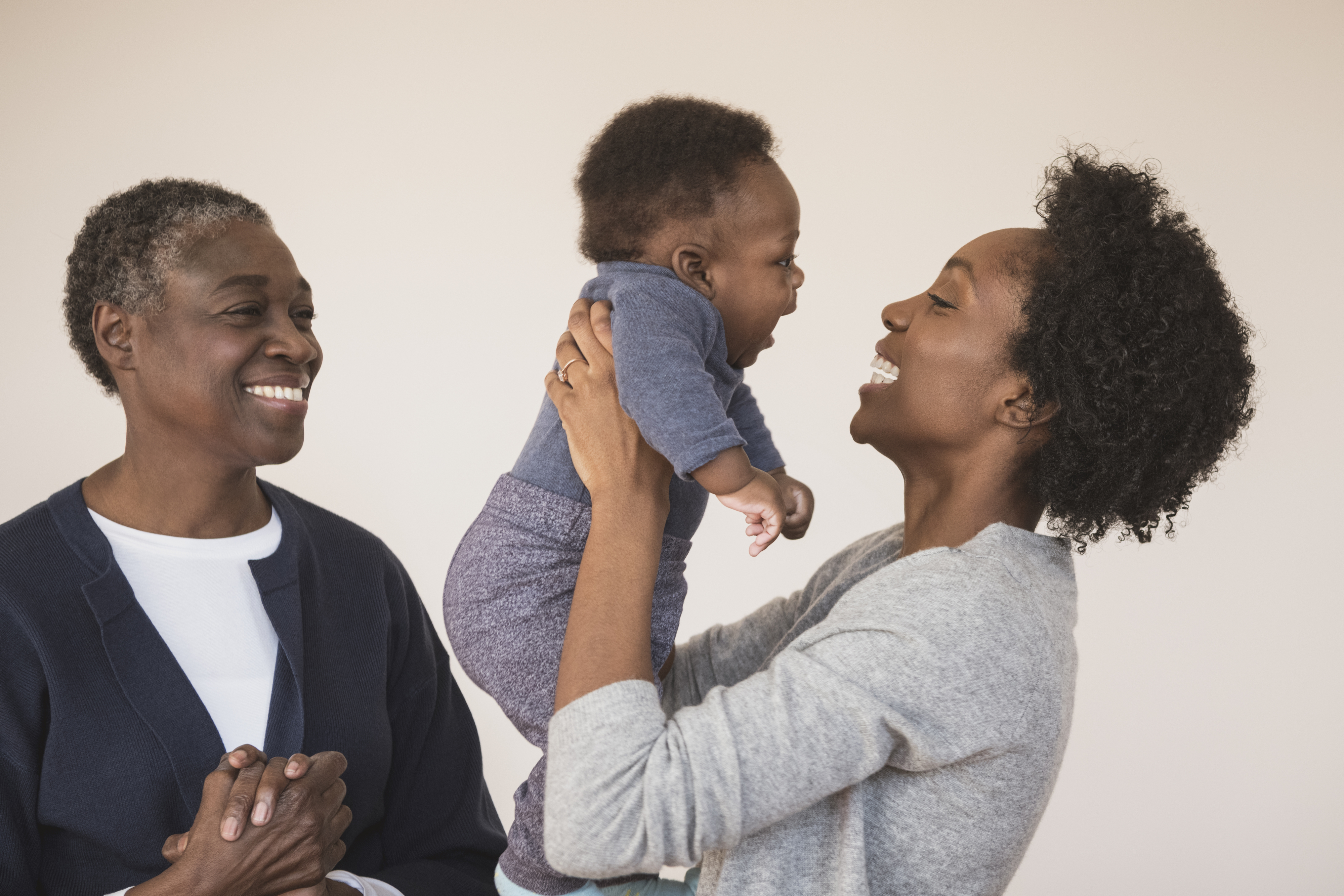 A smiling adult holds a baby playfully, while another adult looks on happily
