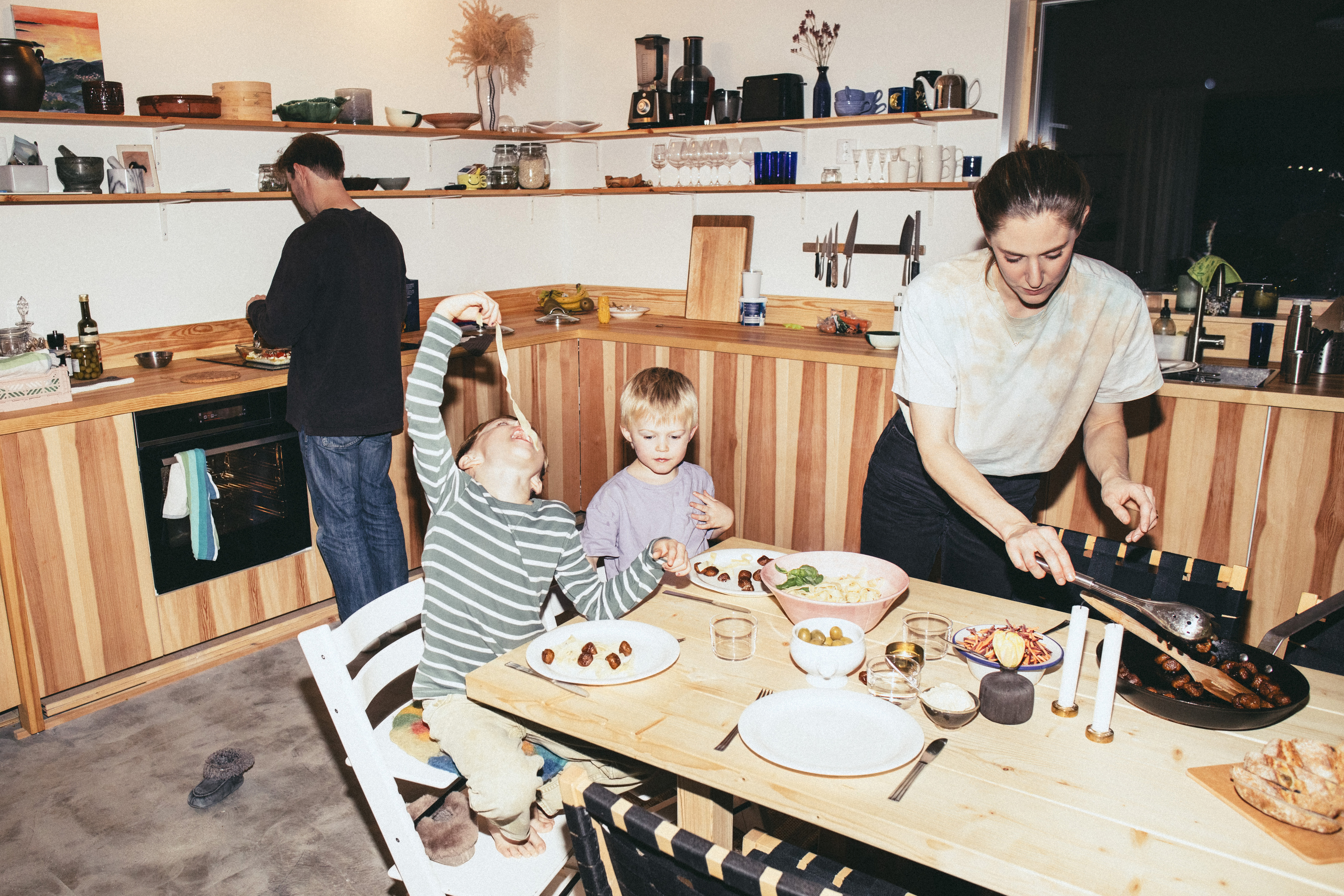A family in a cozy kitchen: a man cooks at the counter while a woman prepares food, and two kids play with table items