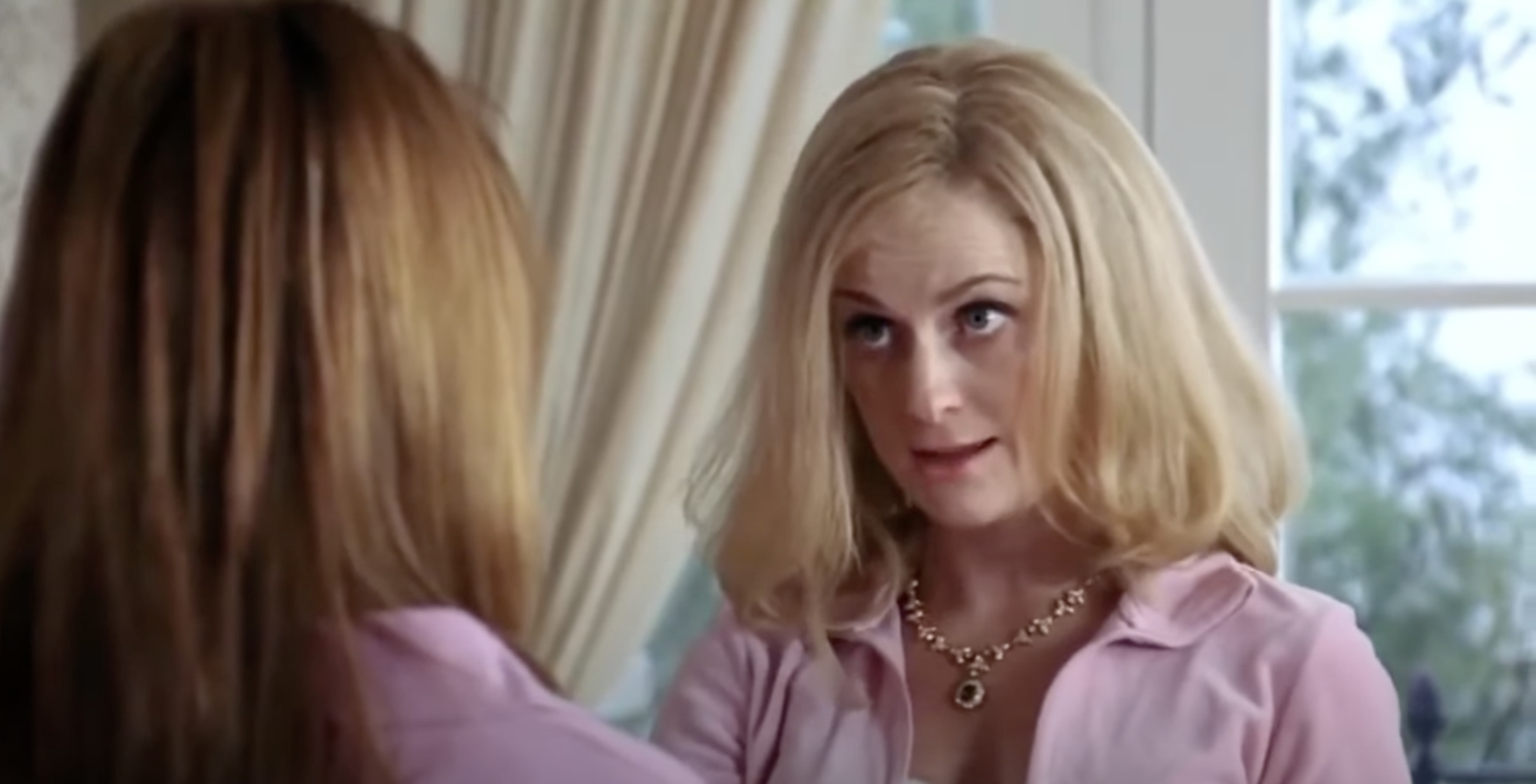From "Mean Girls": Two women in a conversation indoors. One has blonde hair and wears a necklace, and speaks with raised eyebrows to the other