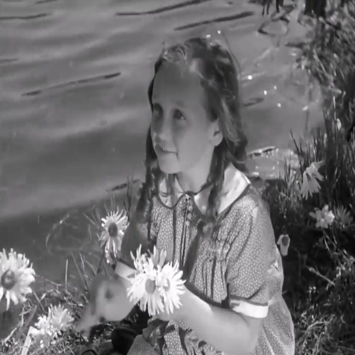 Young girl sitting by a lake, holding flowers, wearing a patterned dress with a white collar, looking upwards