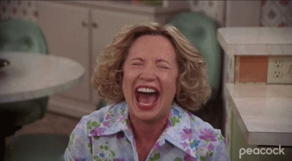 A woman is laughing heartily in a floral blouse in a kitchen setting