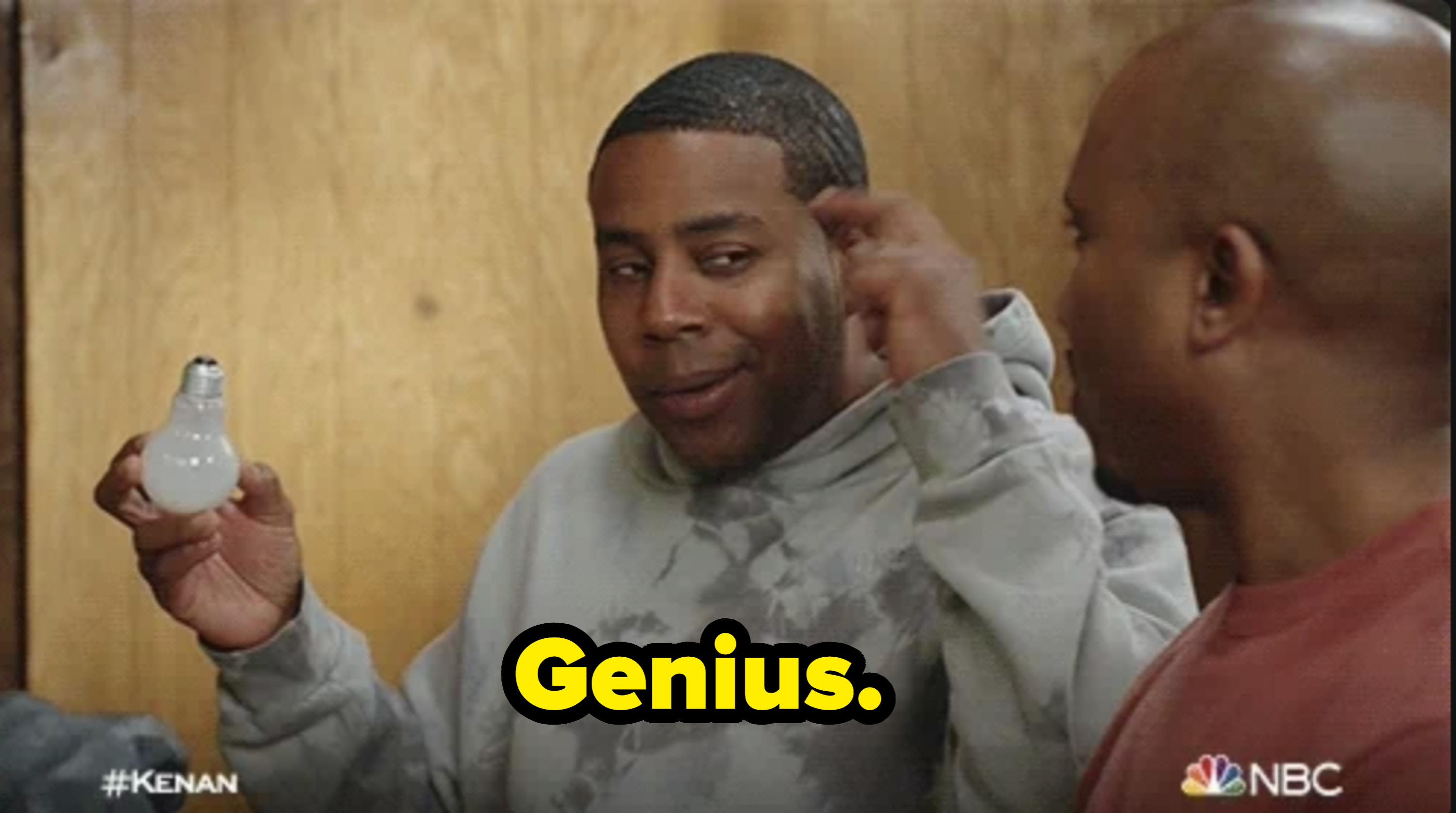 Kenan Thompson smiling, holding a light bulb in a humorous manner, wearing a casual hoodie, with another person listening attentively beside him