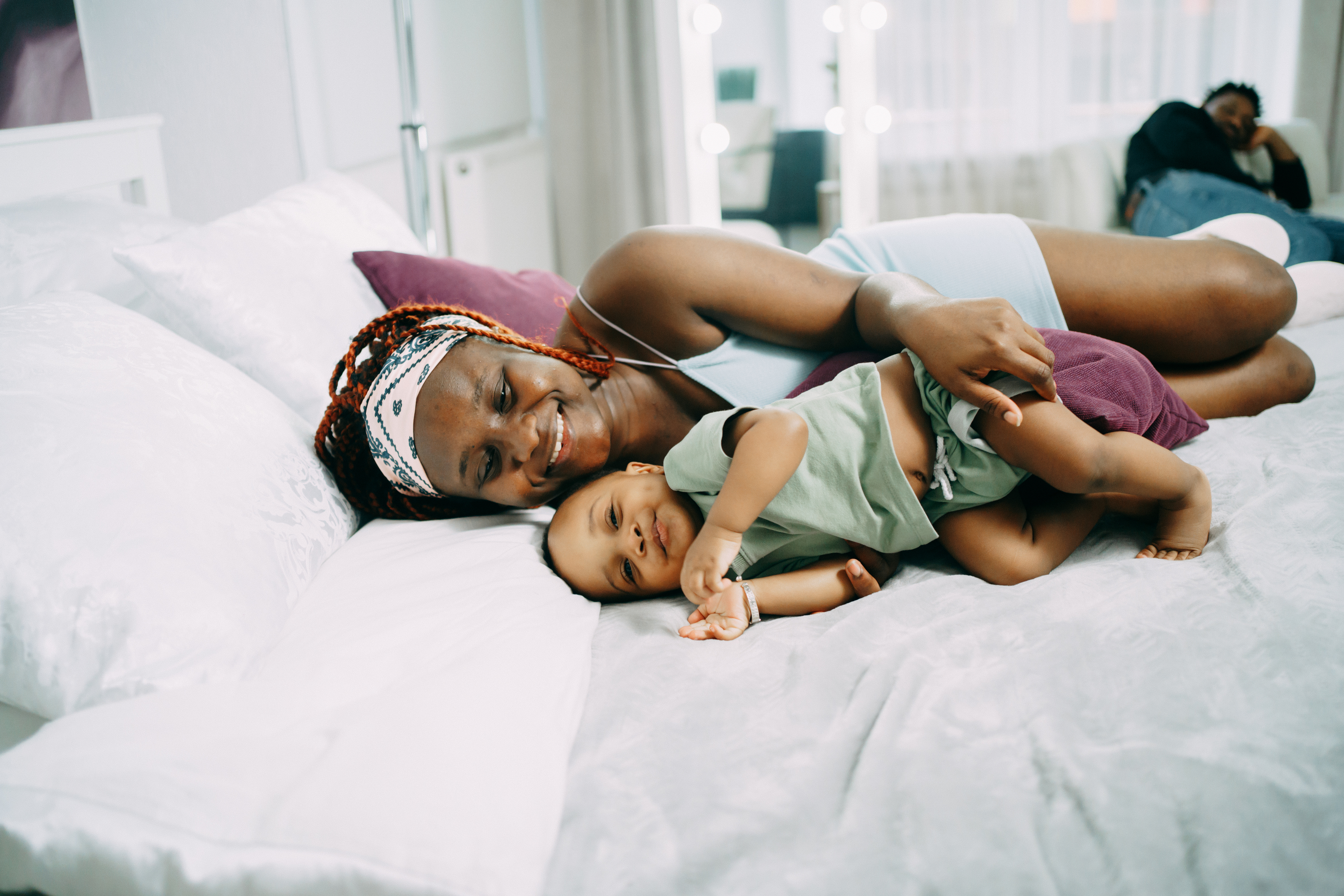 A person relaxes on a bed with a baby, both smiling. Another person is in the background, lounging. Cozy and joyful atmosphere