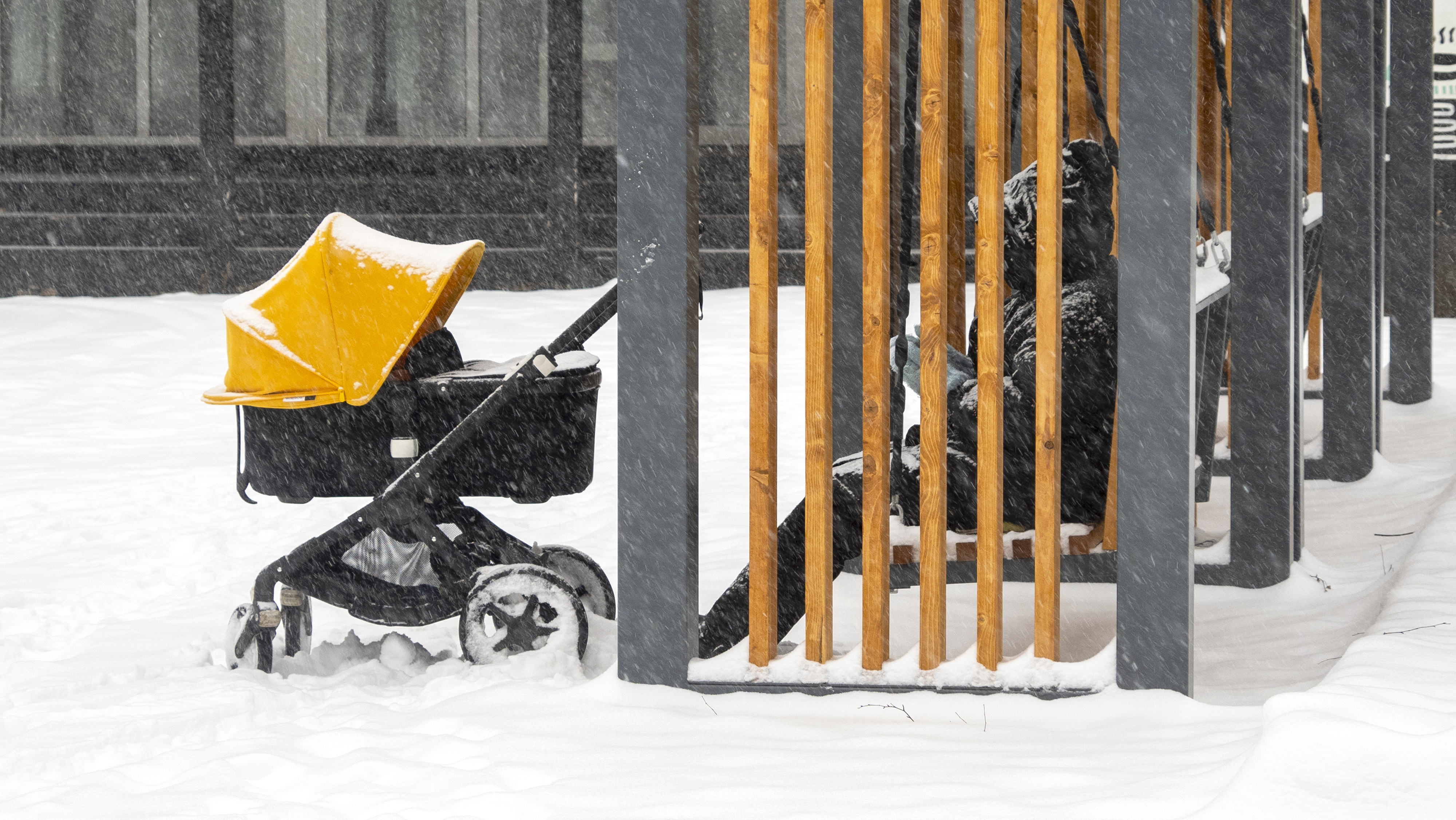Person sitting on a bench in the snow next to a baby stroller, partially covered by wooden slats, on a snowy day