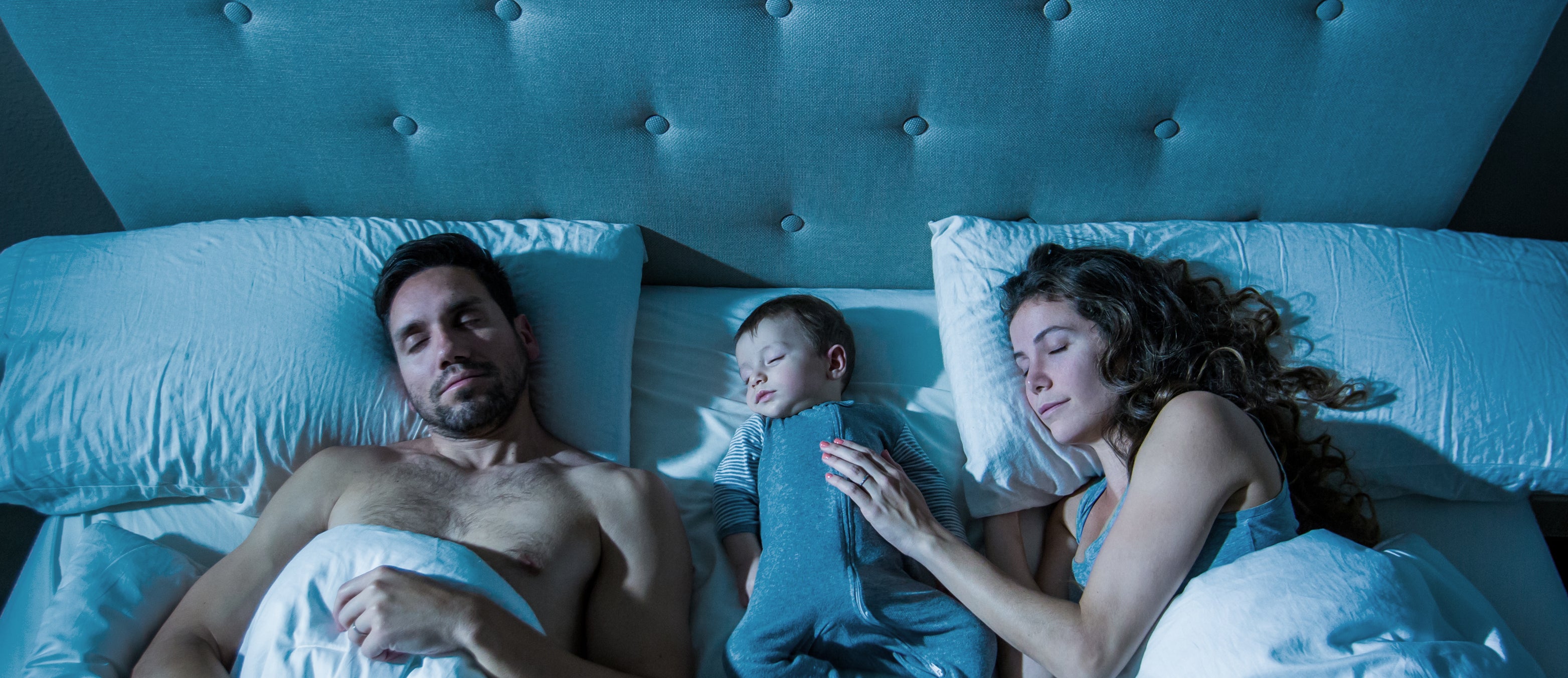 A man, a child, and a woman sleep peacefully in a bed with white sheets and a cushioned headboard