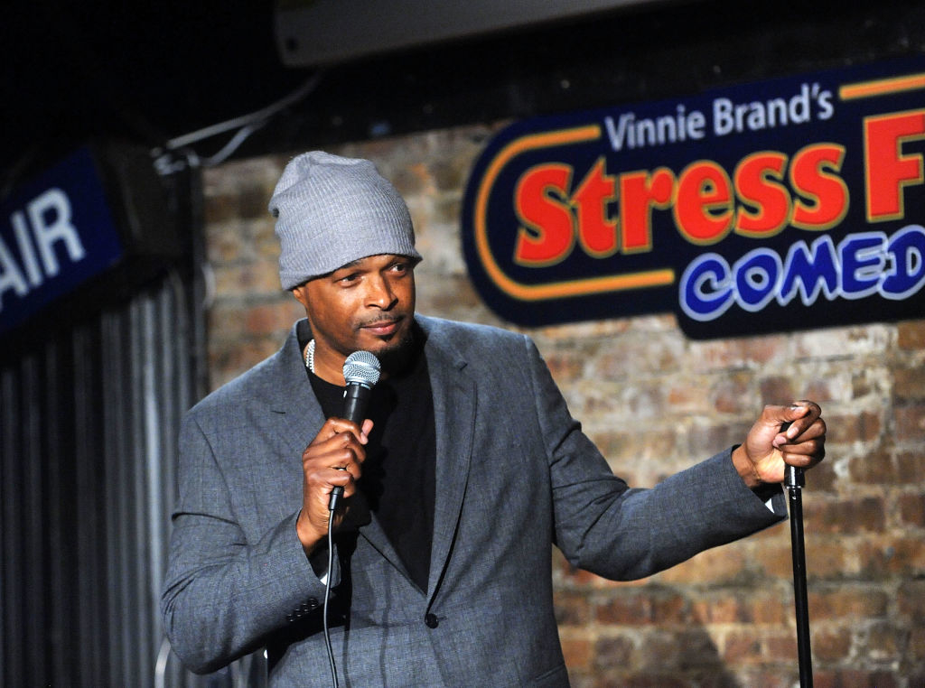 Damon in a gray beanie and suit holds a microphone on stage at Vinnie Brand&#x27;s Stress Factory Comedy Club