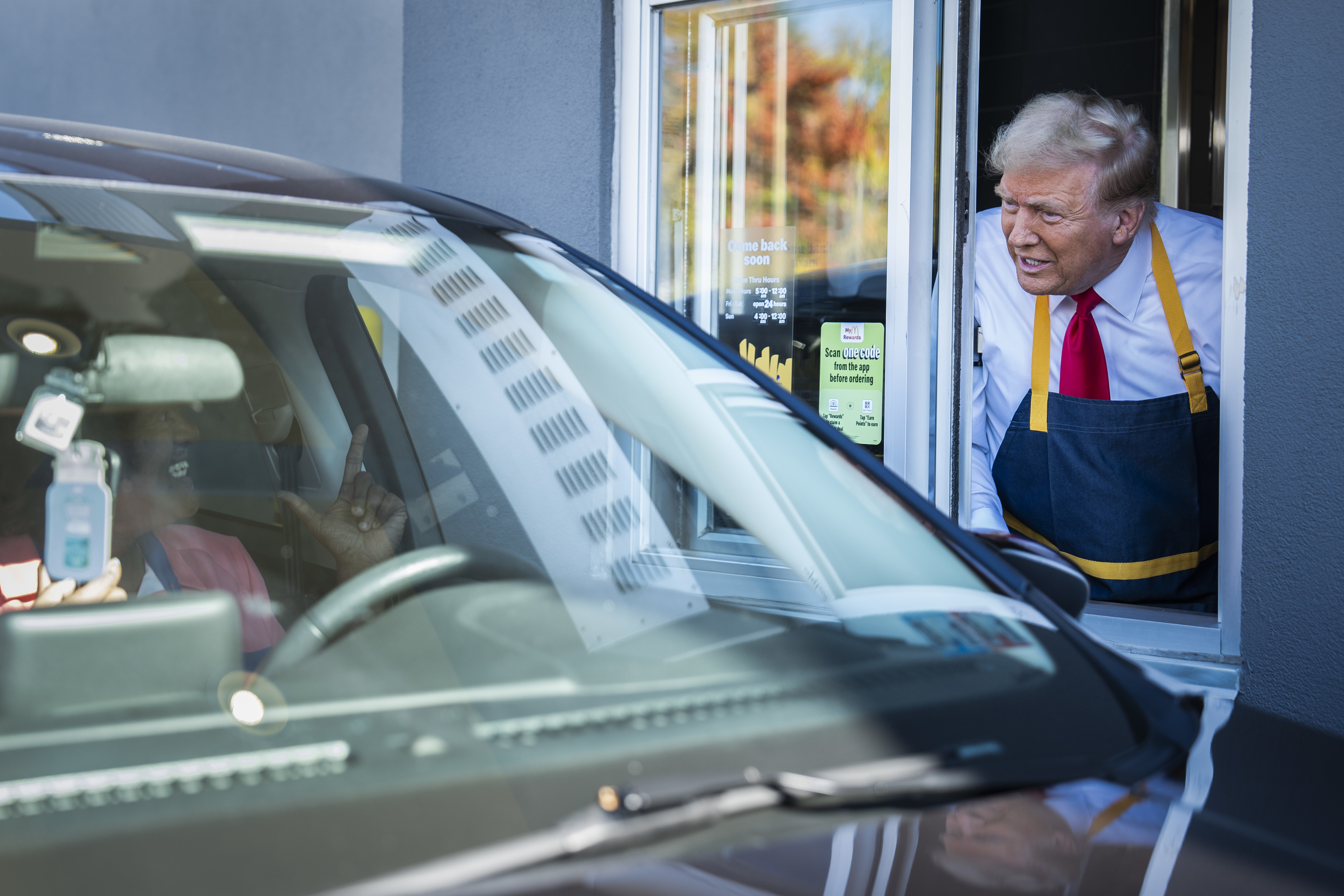 I don&#x27;t know who is in this image, but it shows a drive-thru worker wearing a blue apron and white shirt speaking to a driver in a car