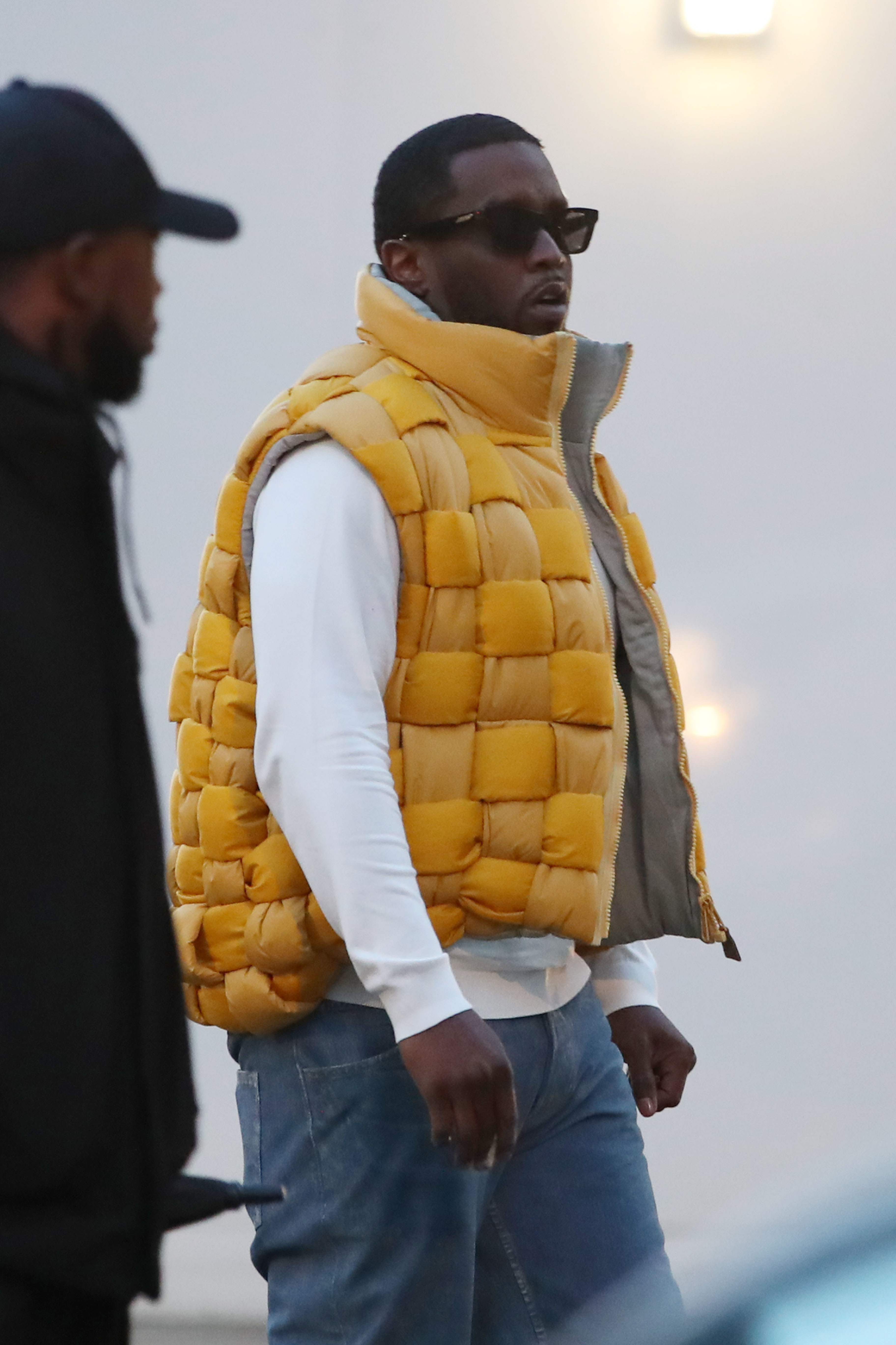 A person in a puffy vest, sunglasses, and jeans walks outdoors