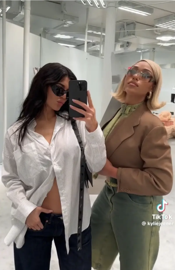 Two women posing for a mirror selfie. The woman on the left wears a partially open shirt and sunglasses. The woman on the right wears a blazer and glasses