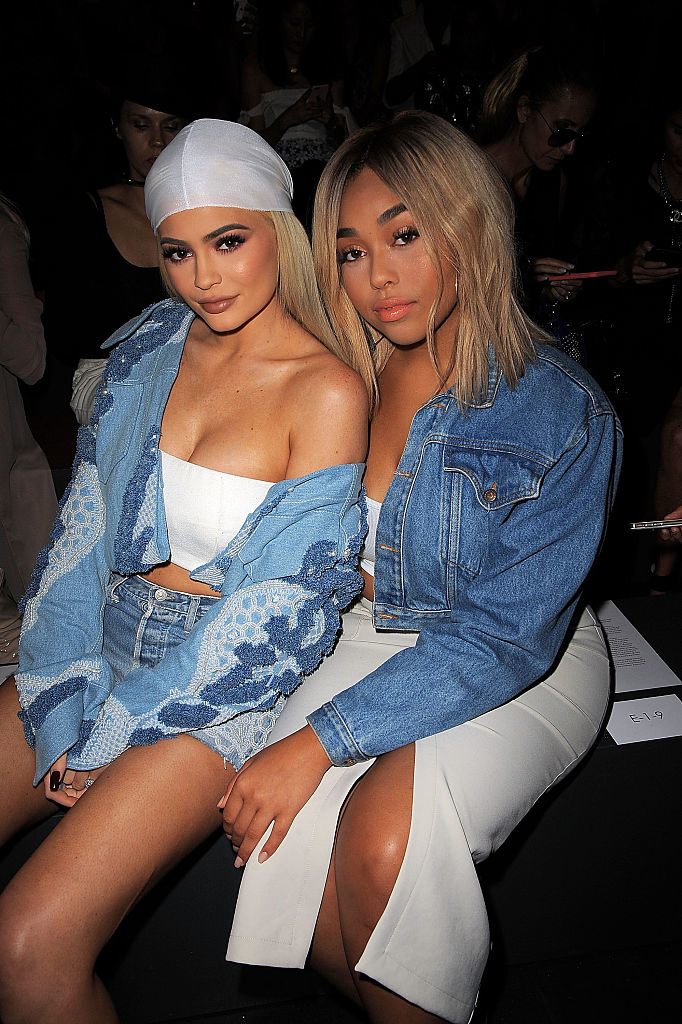 Two women sitting together, wearing denim jackets over white outfits, posing for a photo at a celebrity event