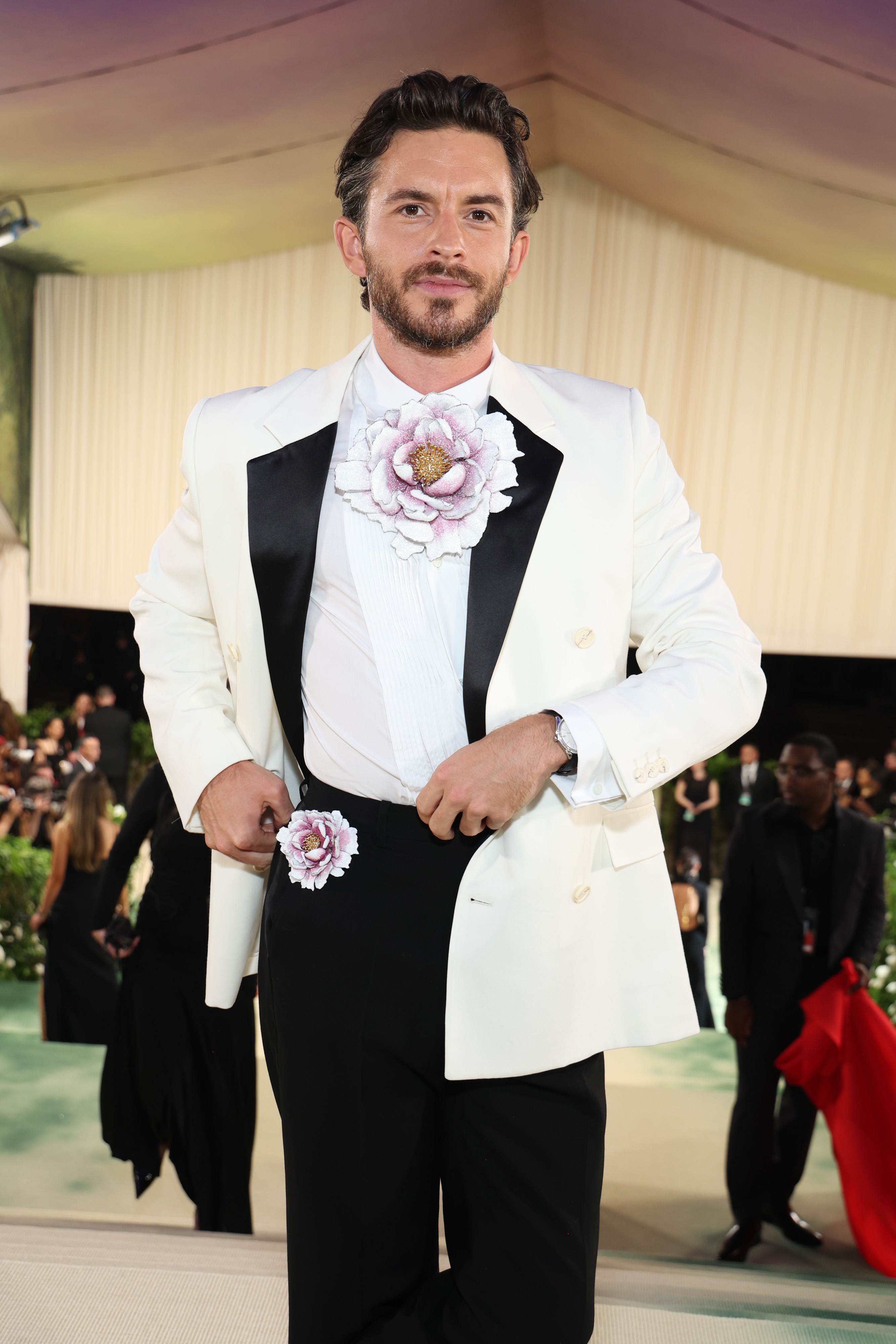 A man in a white tuxedo jacket with large floral detail on the lapel stands on a red carpet