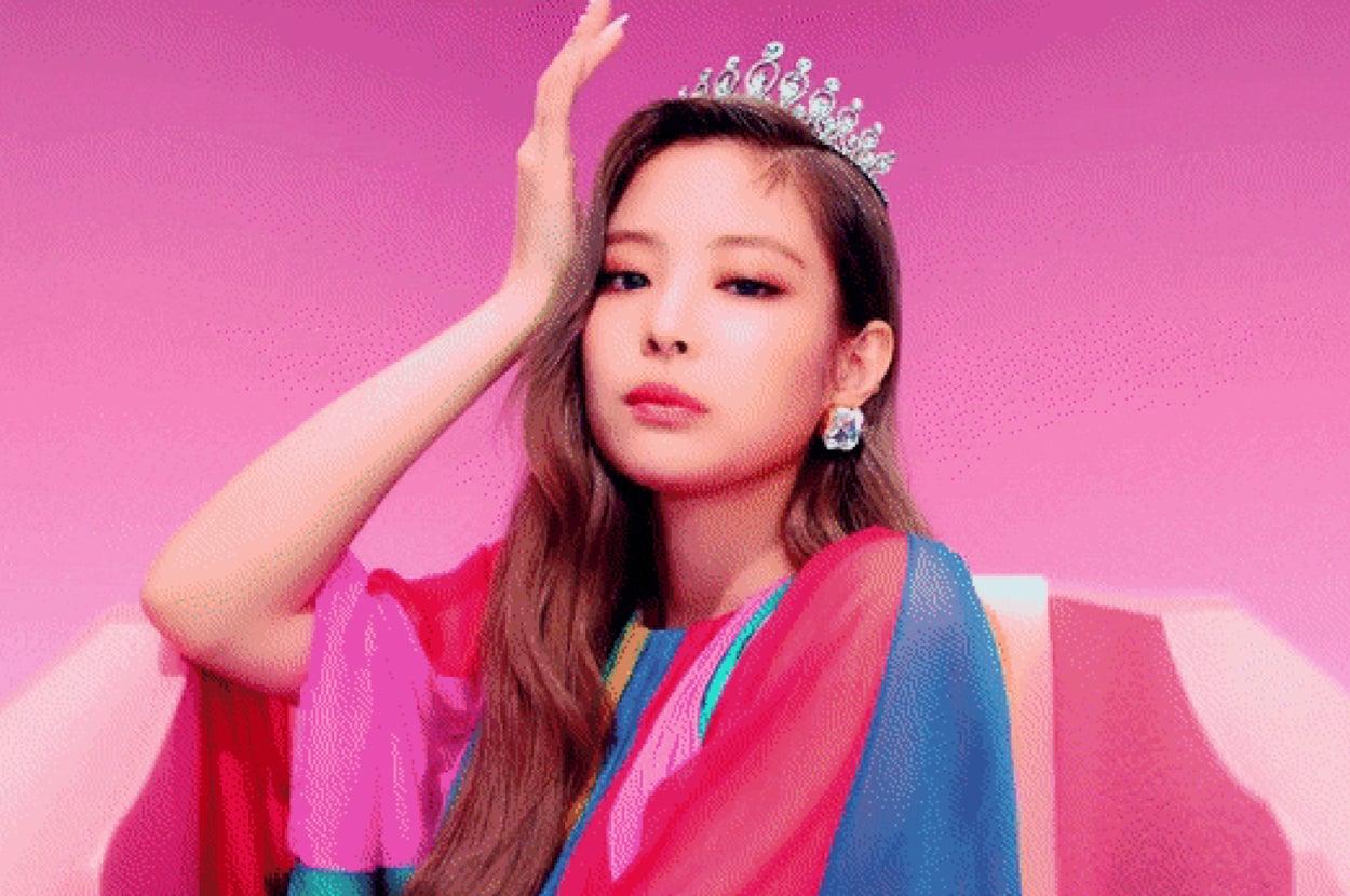 Jenny poses with a hand on their head, wearing a tiara and colorful outfit, sitting against a vibrant backdrop