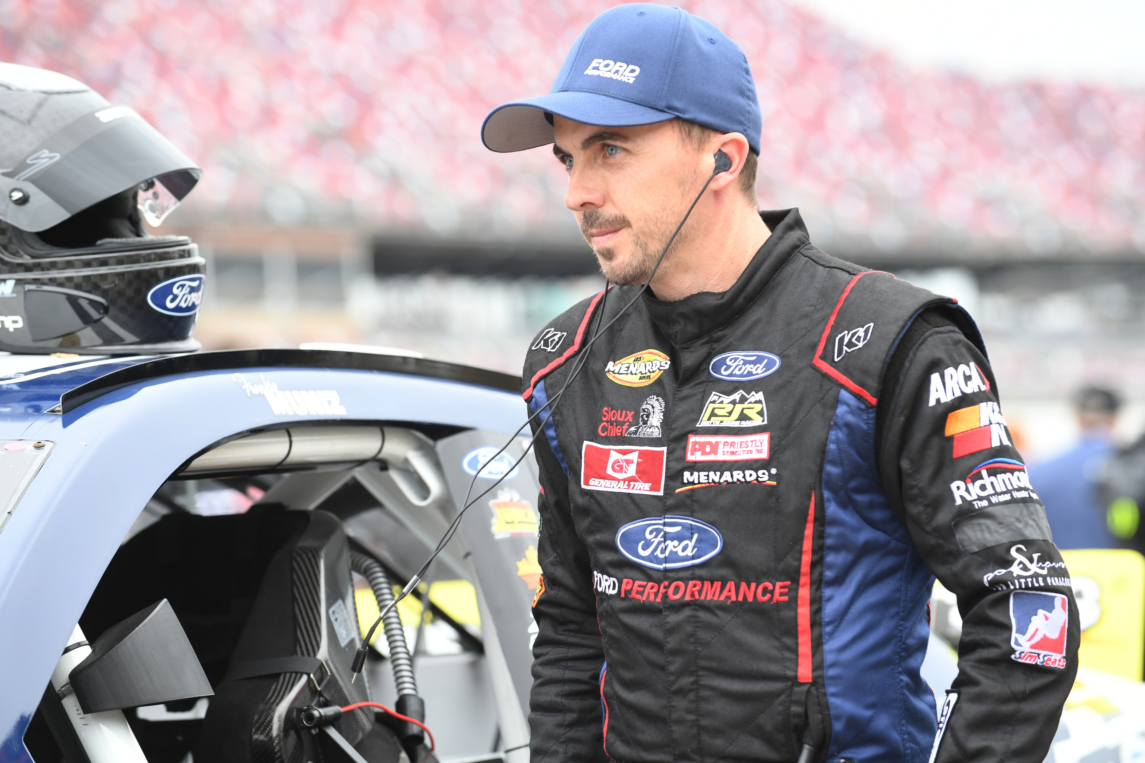 A racecar driver in a Ford jumpsuit and blue cap stands beside a car, wearing an earpiece, with a focused expression