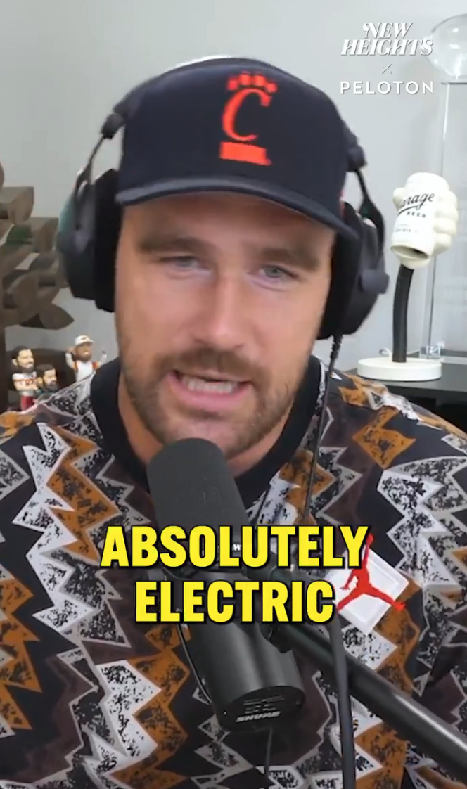 Person wearing a patterned sweater and cap, speaking into a microphone with bold text reading &quot;ABSOLUTELY ELECTRIC&quot; on the image