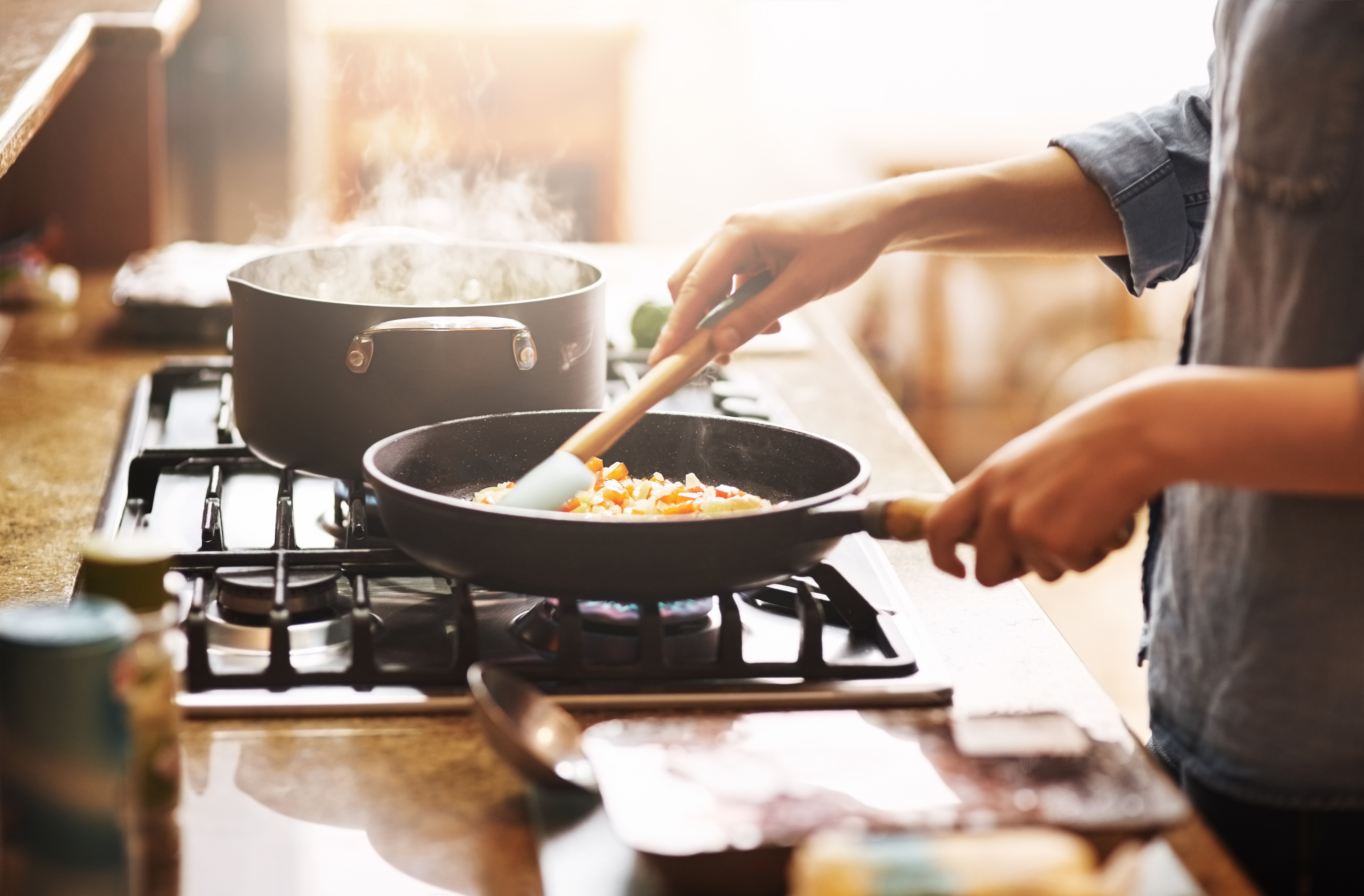 Person cooking on a stovetop, stirring vegetables in a pan, with a pot of boiling water nearby