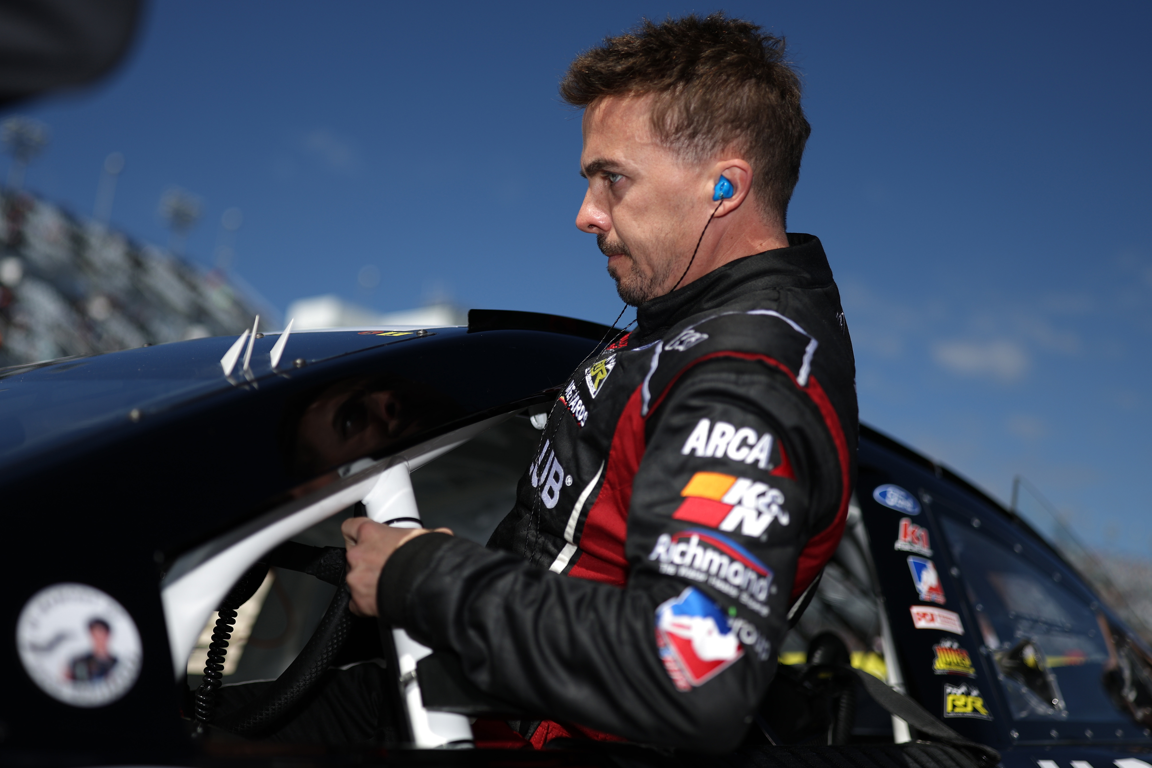 A person in a racing suit with sponsor logos wearing earplugs looks focused while preparing for a race in a car