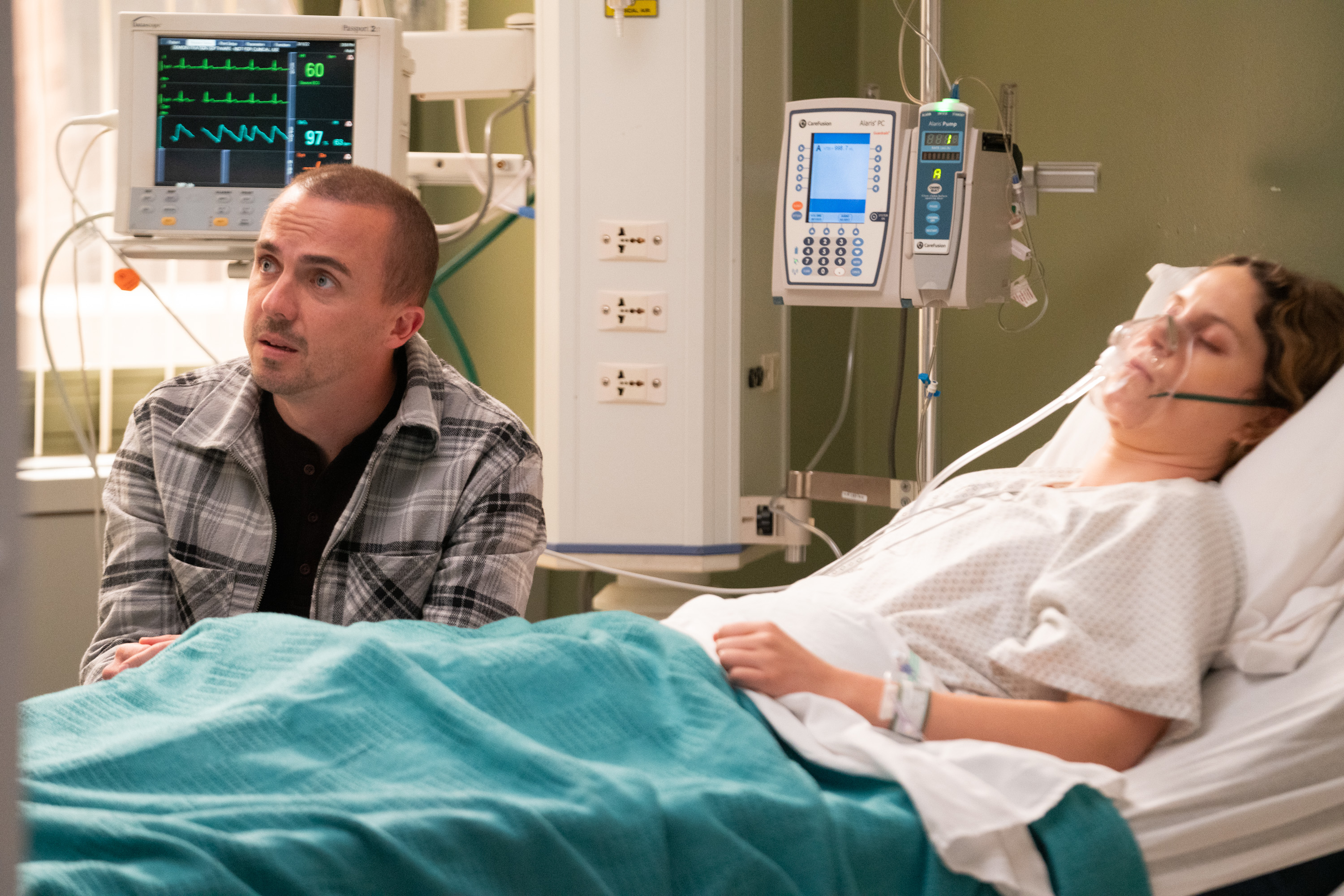 A man sits next to a woman in a hospital bed, wearing a gown and oxygen mask, surrounded by medical equipment