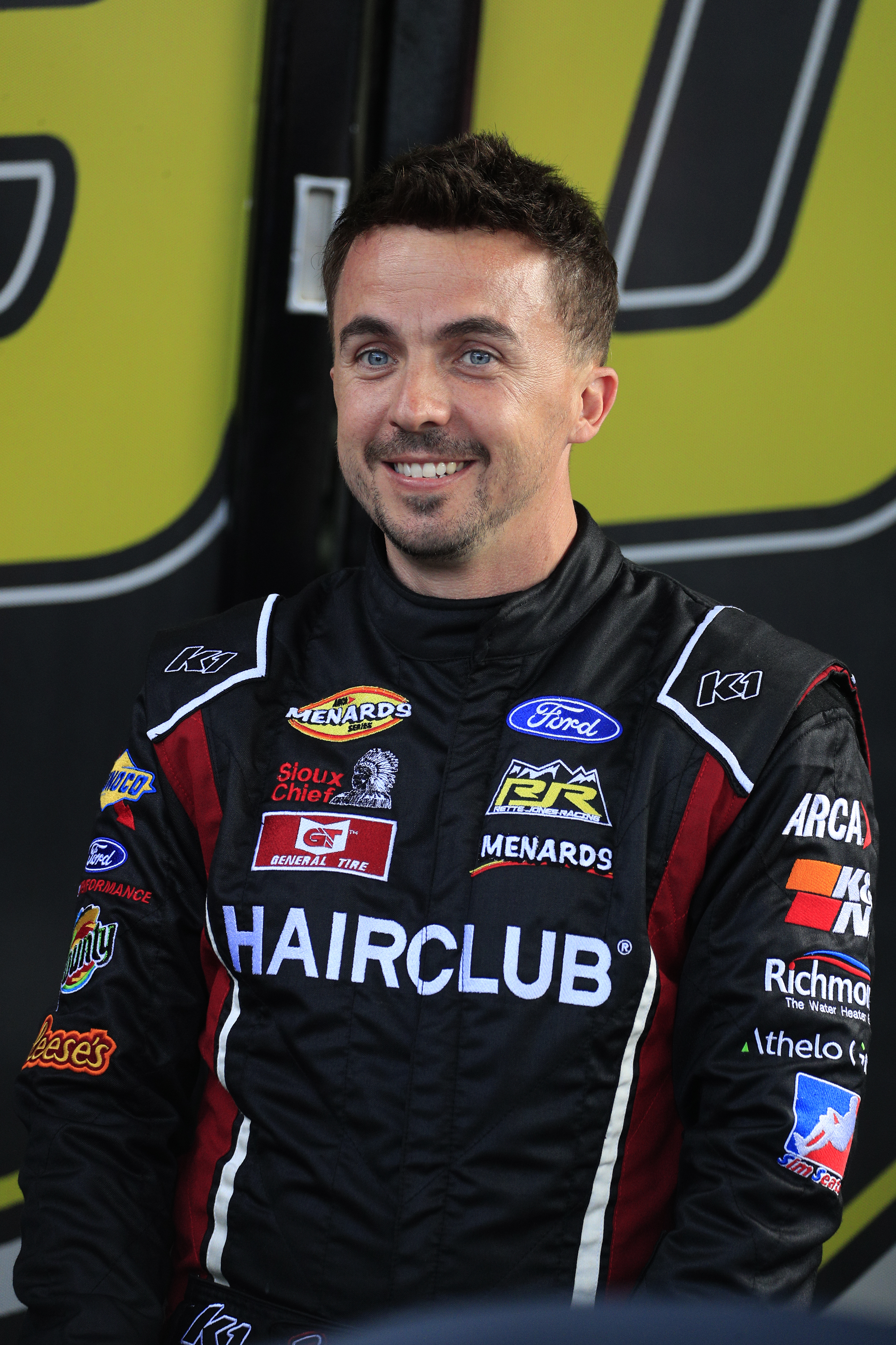 A man in a racing suit with various sponsor logos, smiling