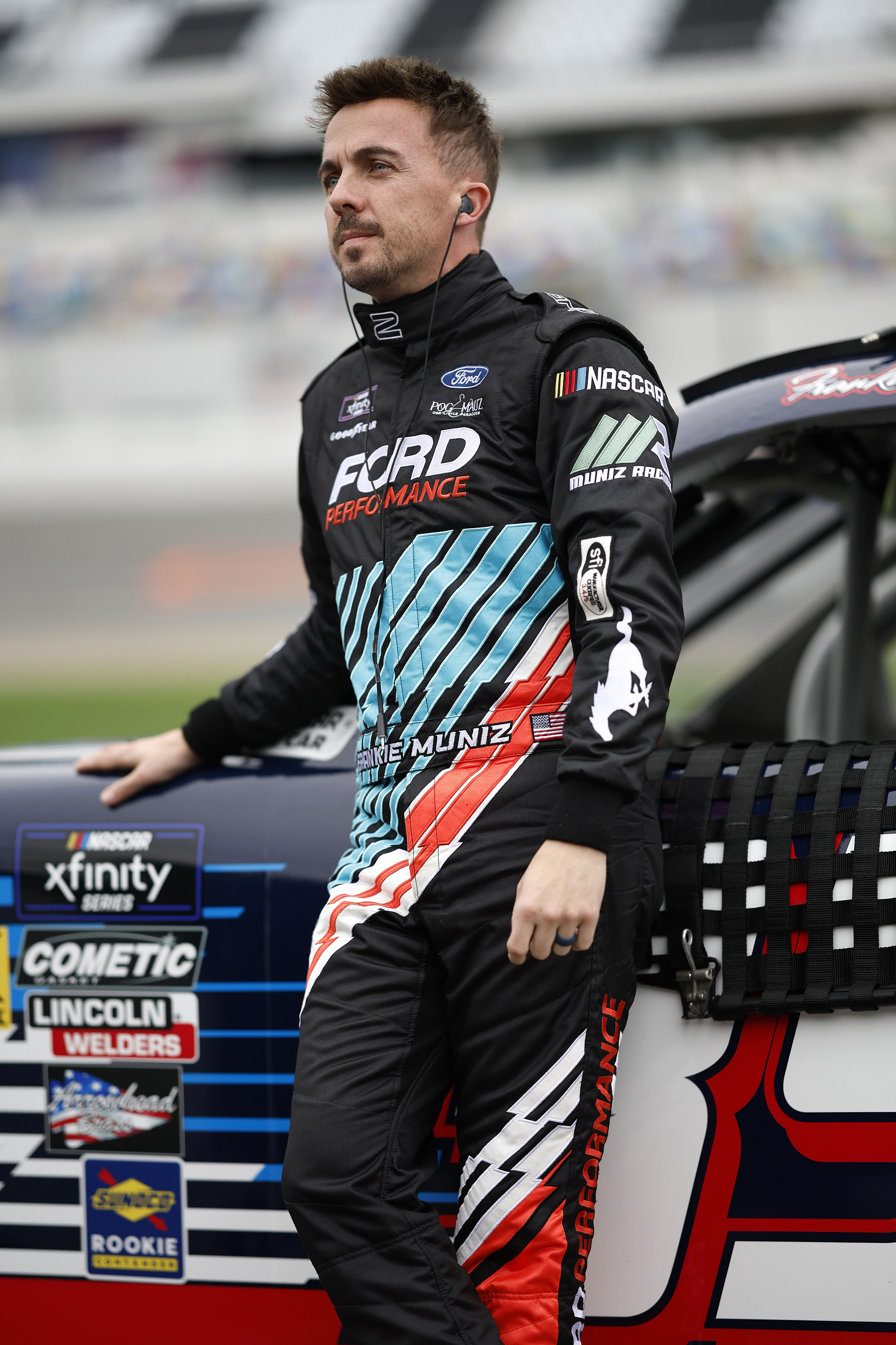 A racecar driver in a Ford Performance suit stands beside a racecar, focused and poised