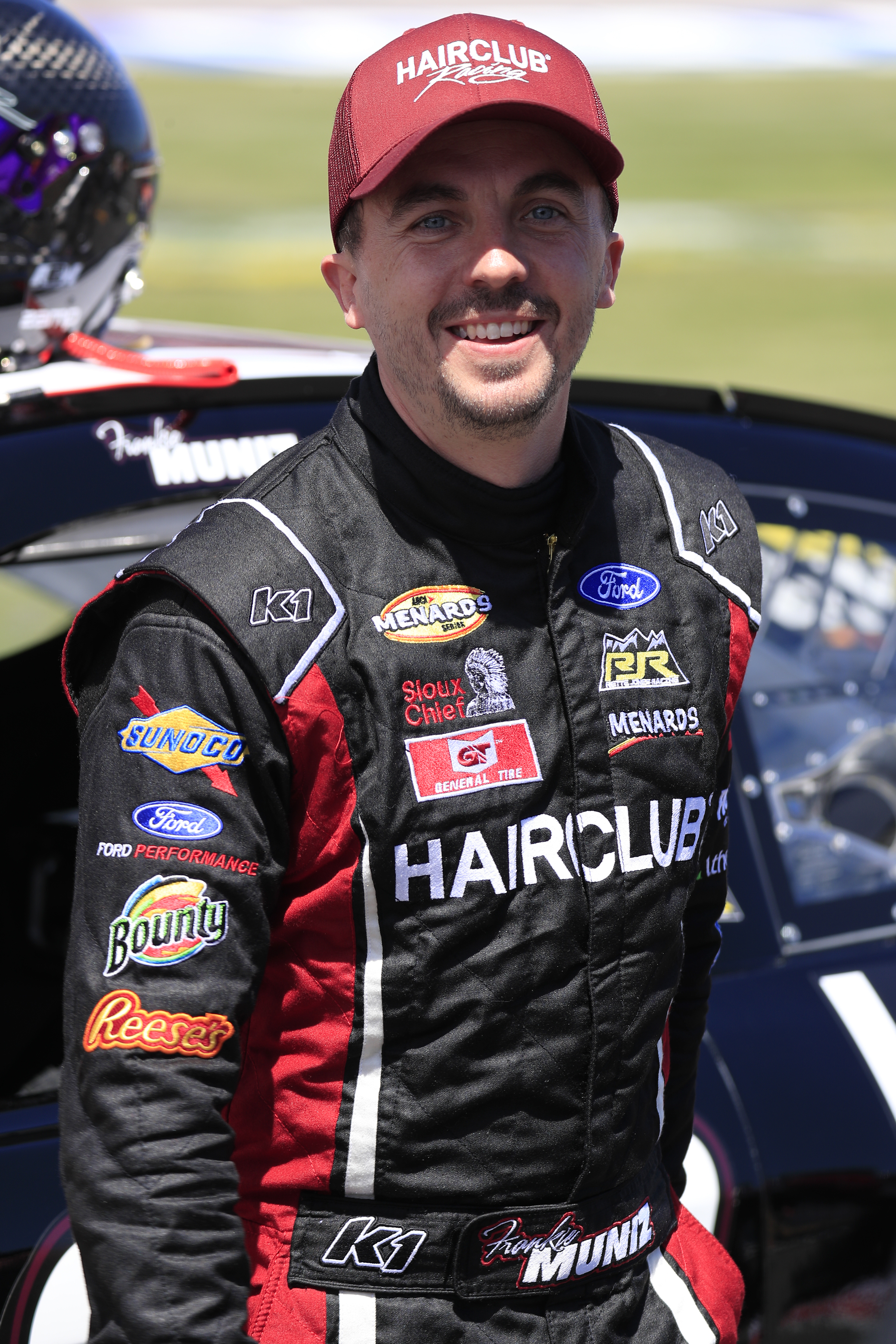 A person in a racing suit and cap stands by a race car, smiling. Various sponsor logos are visible on the suit