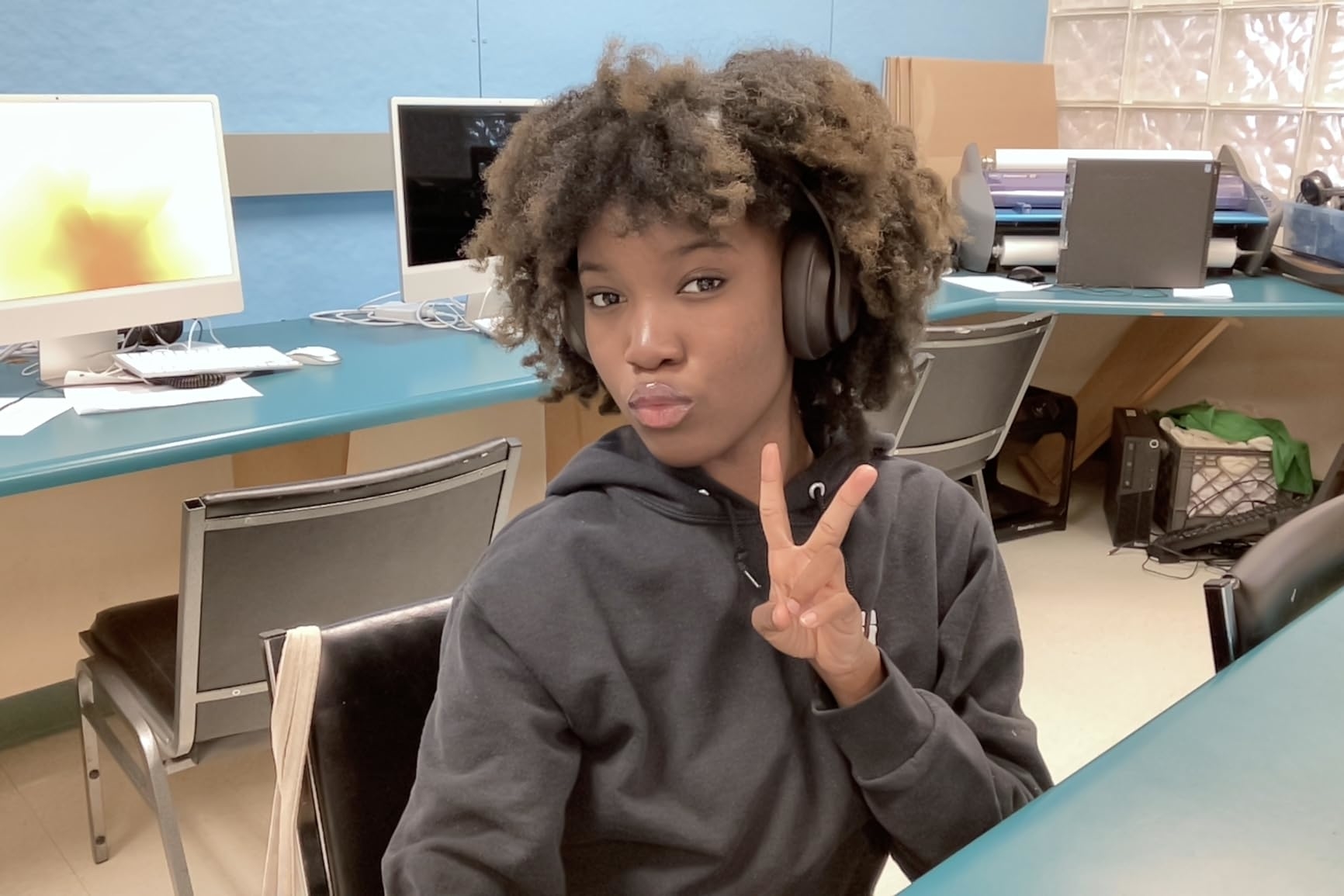 Person poses with headphones, makes a peace sign in a tech workspace with computers.