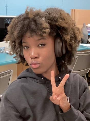 Person poses with headphones, makes a peace sign in a tech workspace with computers.
