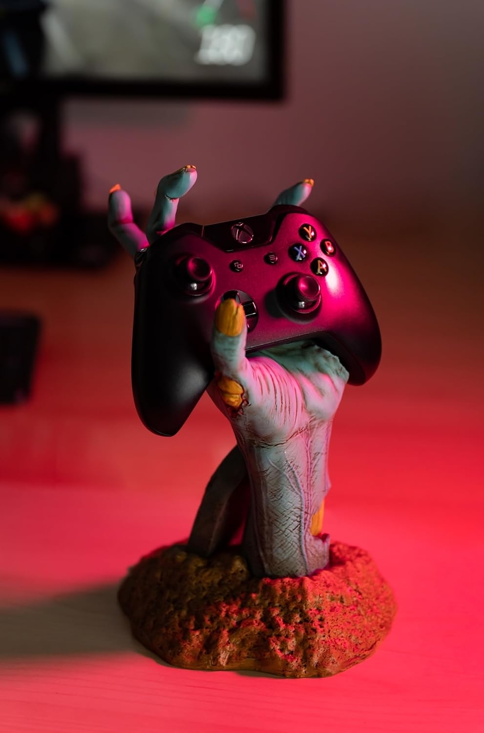 A decorative zombie hand holding a gaming controller, used as a stand, placed on a desk with a gaming setup in the background