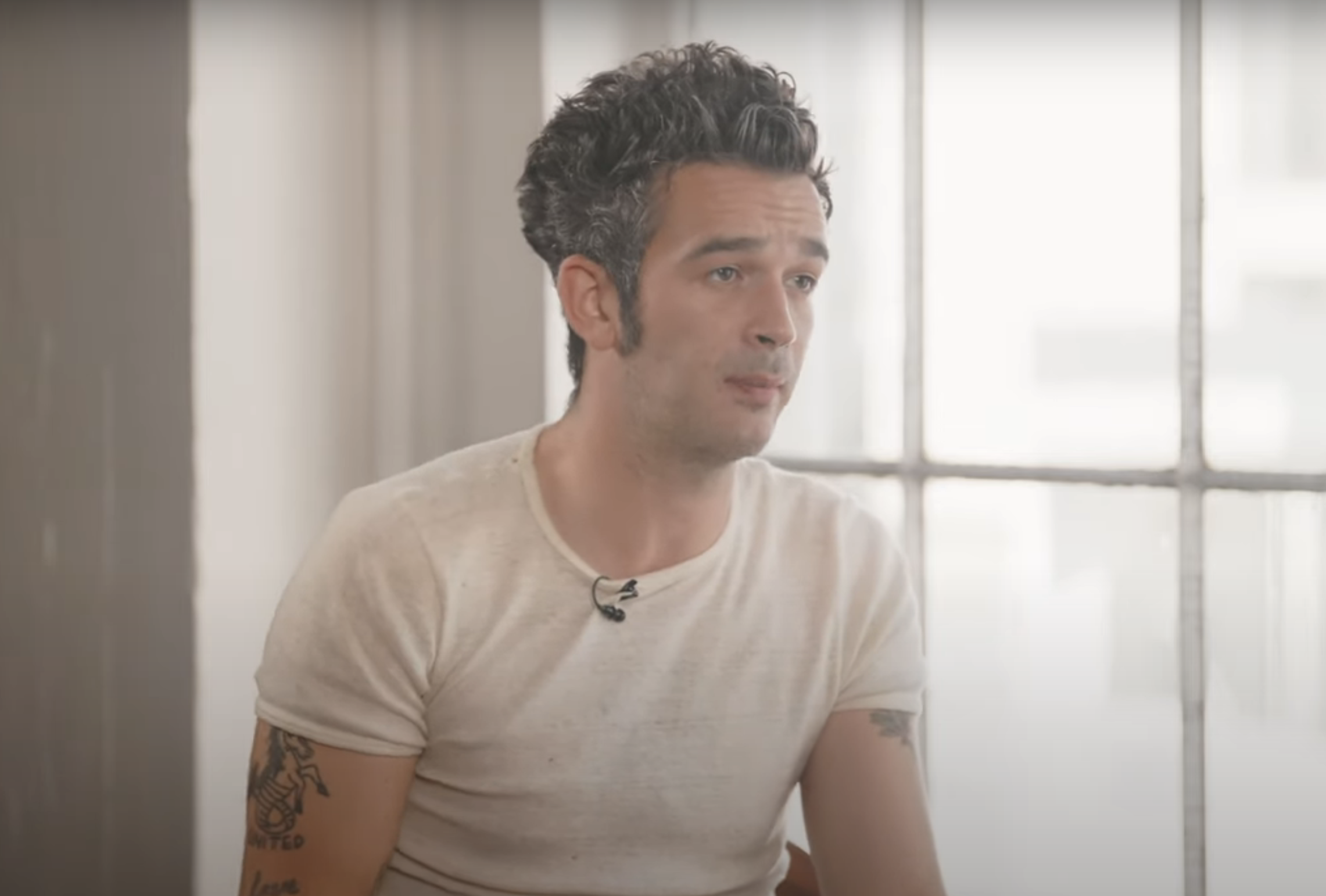 Matty Healy with styled hair and visible tattoos is sitting in front of a window, wearing a light-colored t-shirt and speaking in a candid setting