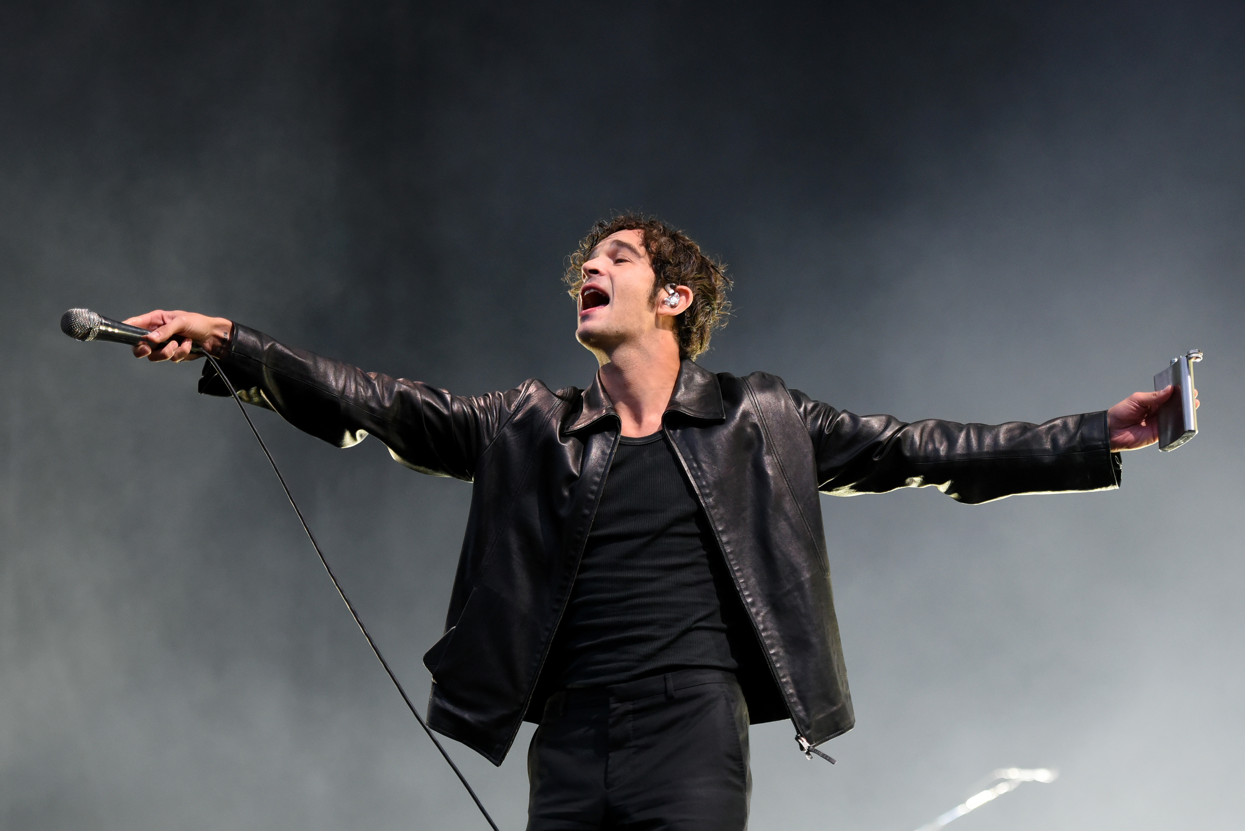 Matty Healy in a  leather jacket energetically performs on stage, holding a microphone in one hand and a small object in the other