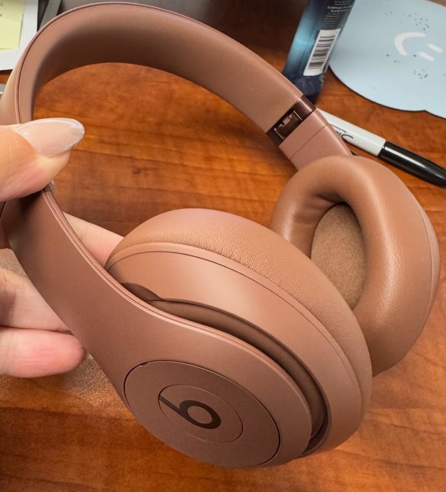 Brown Beats headphones held in a person's hand, displayed on a wooden surface with a pen and a light blue mouse pad in the background
