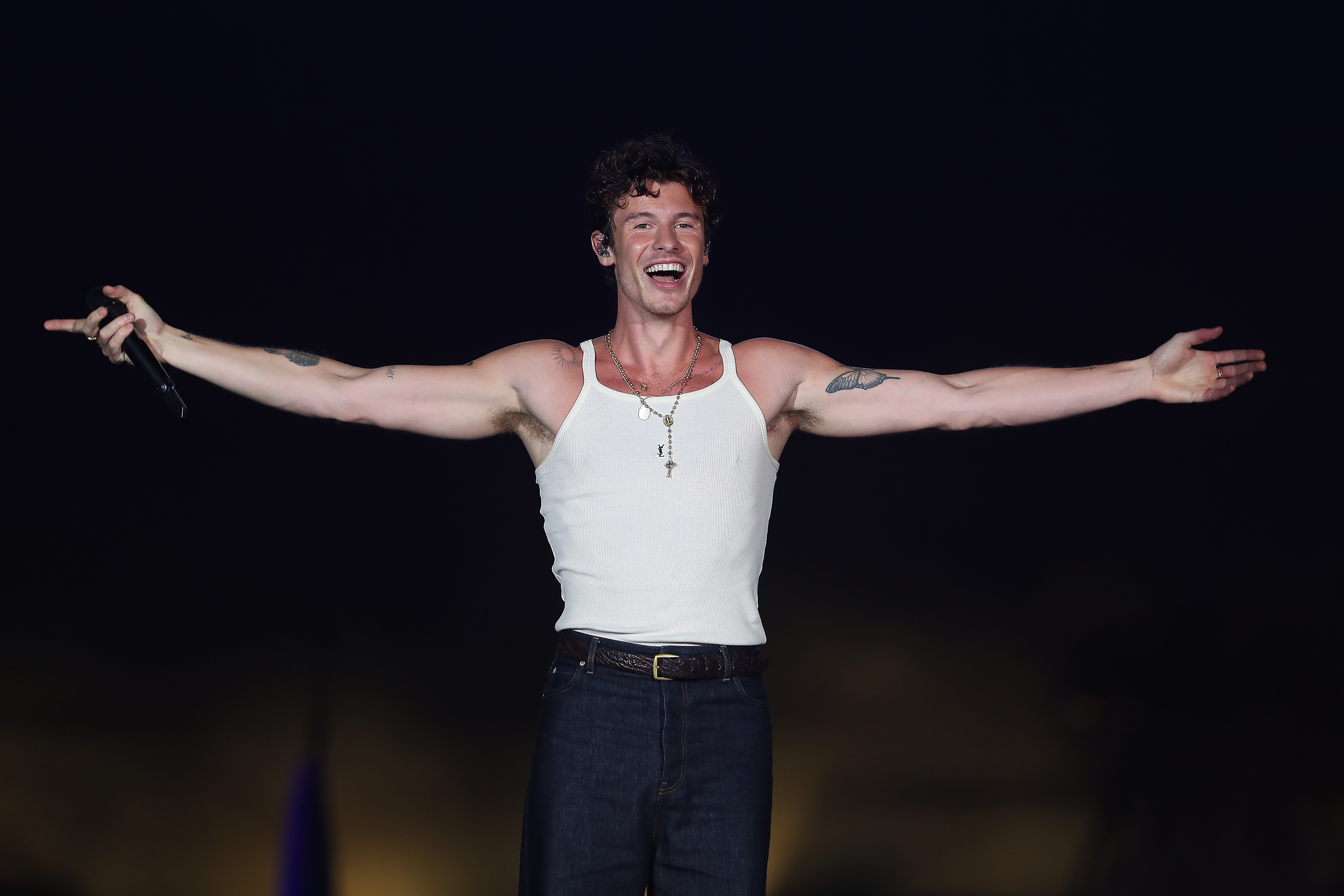 A man wearing a white tank top and jeans performs on stage, smiling with arms outstretched, holding a microphone