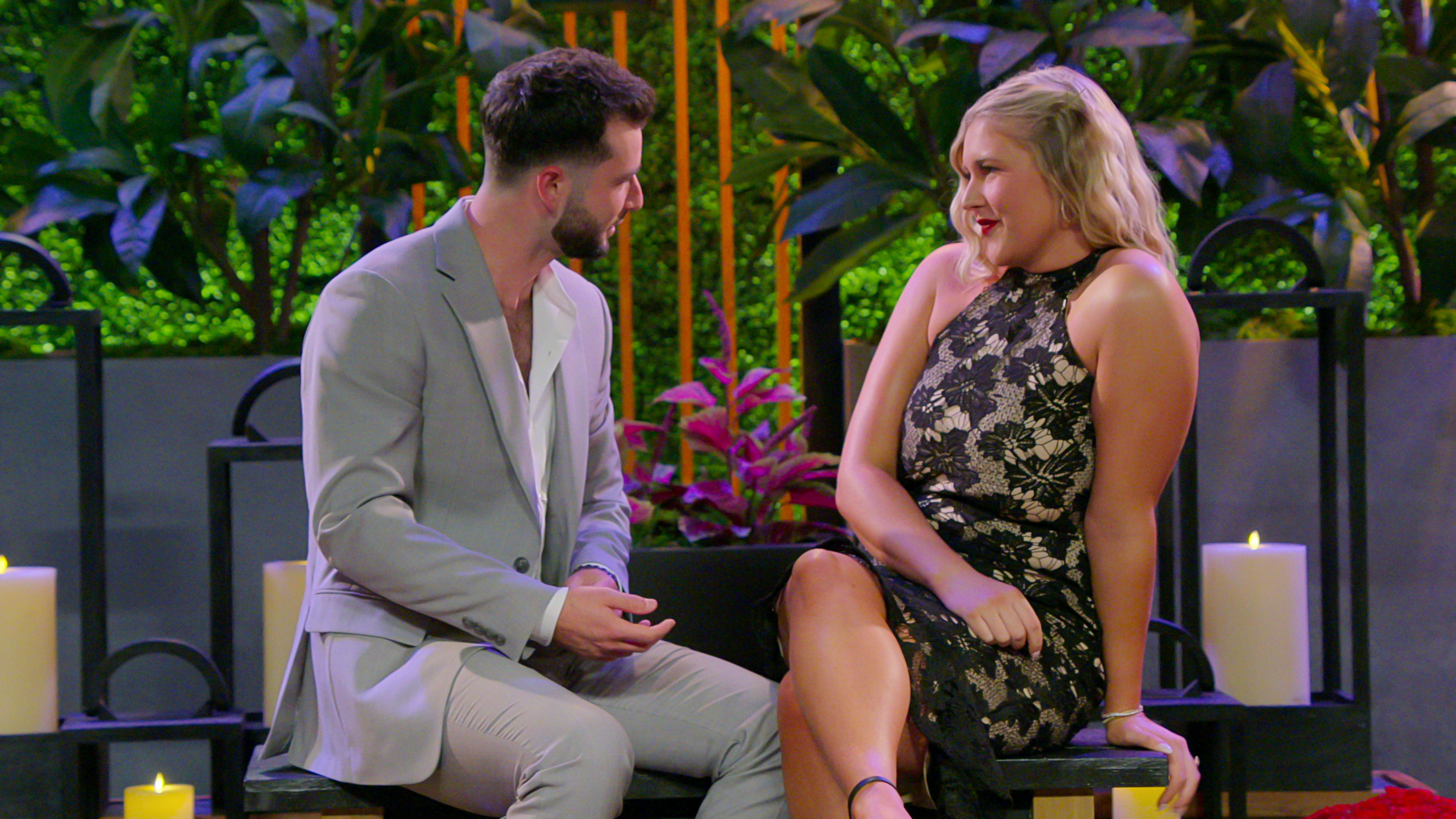 Man in light suit and woman in floral dress sit together on a set with plants and candles in the background, engaged in conversation