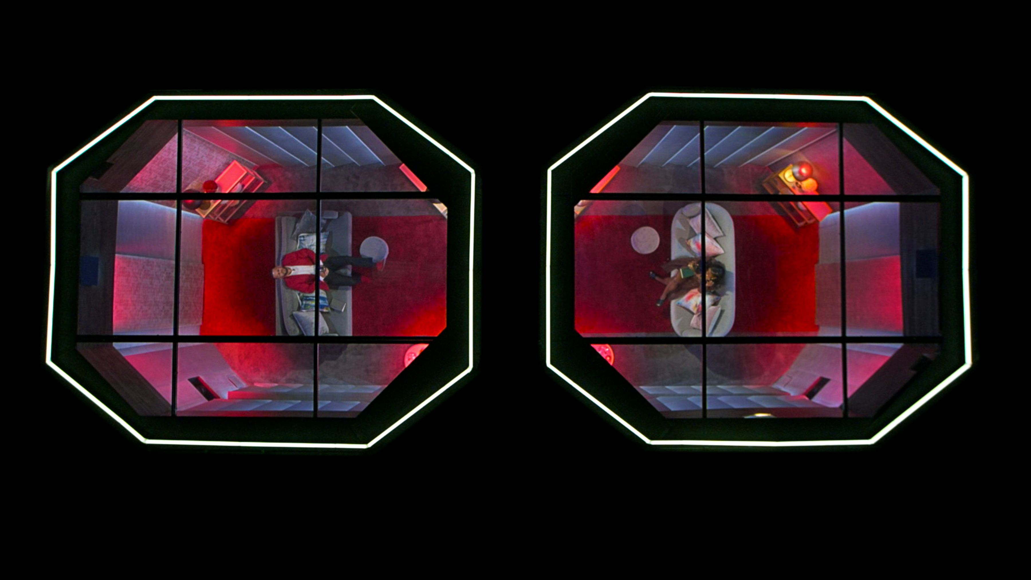 Overhead view of two octagonal glass enclosures, each with one person lying inside, surrounded by red lighting