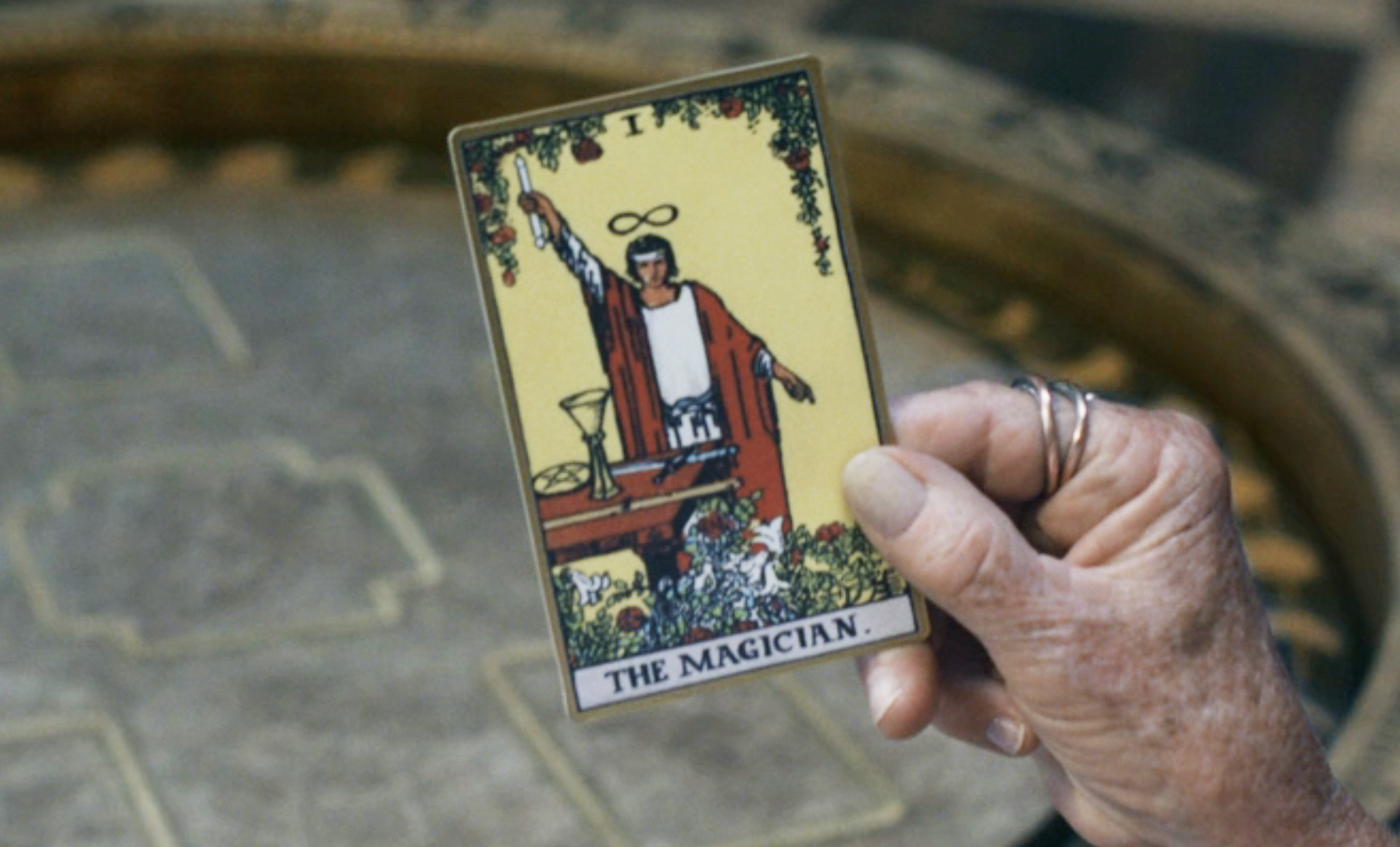 Hand holding a tarot card labeled &quot;The Magician,&quot; depicting a figure with a raised hand and a table with items, in a mystic setting