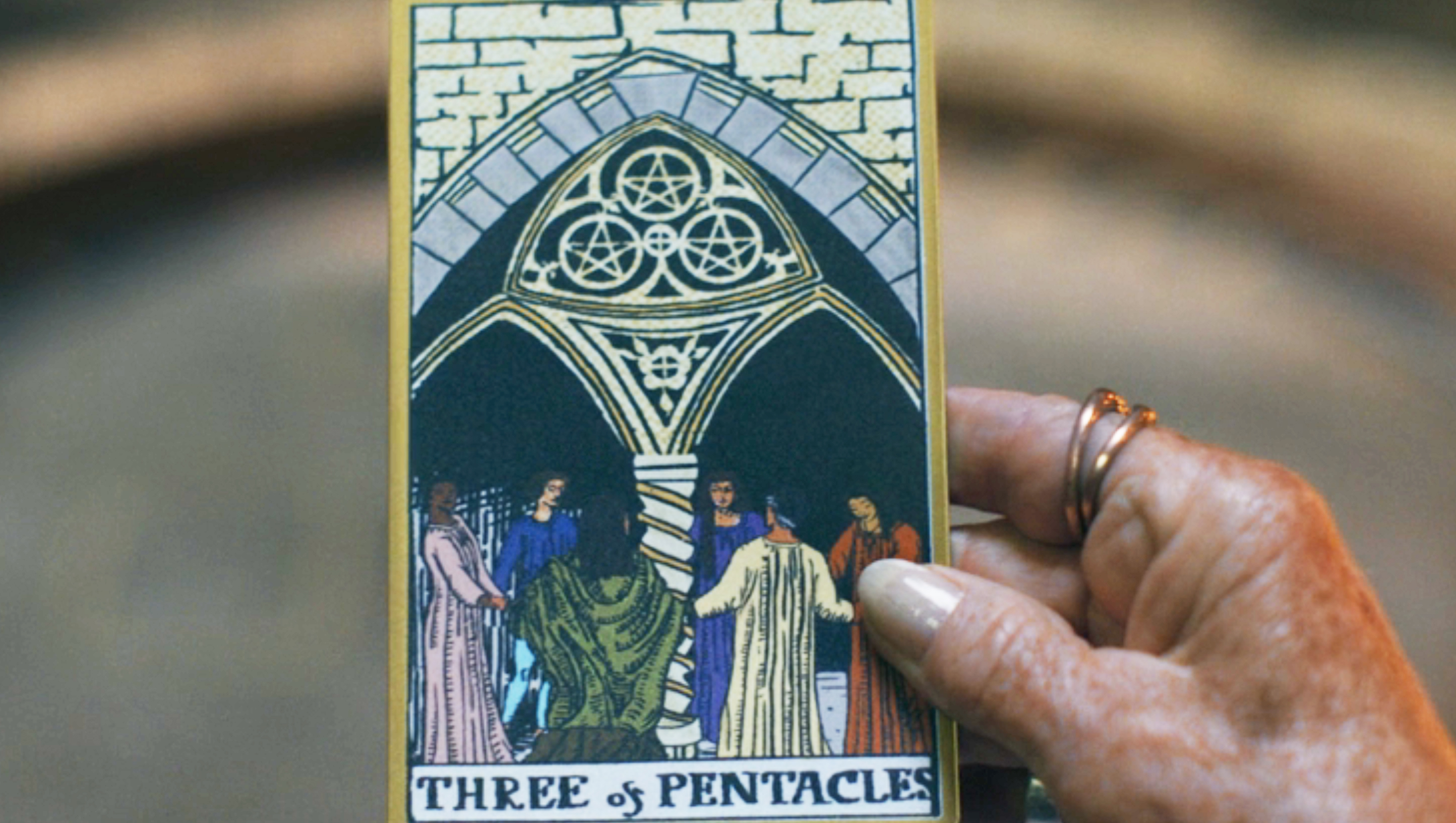 A close up of a hand holding a tarot card depicting the Three of Pentacles