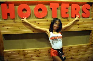 A person posing with arms outstretched in front of a wooden wall with a large red "Hooters" sign, wearing a Hooters uniform with a graphic tank top
