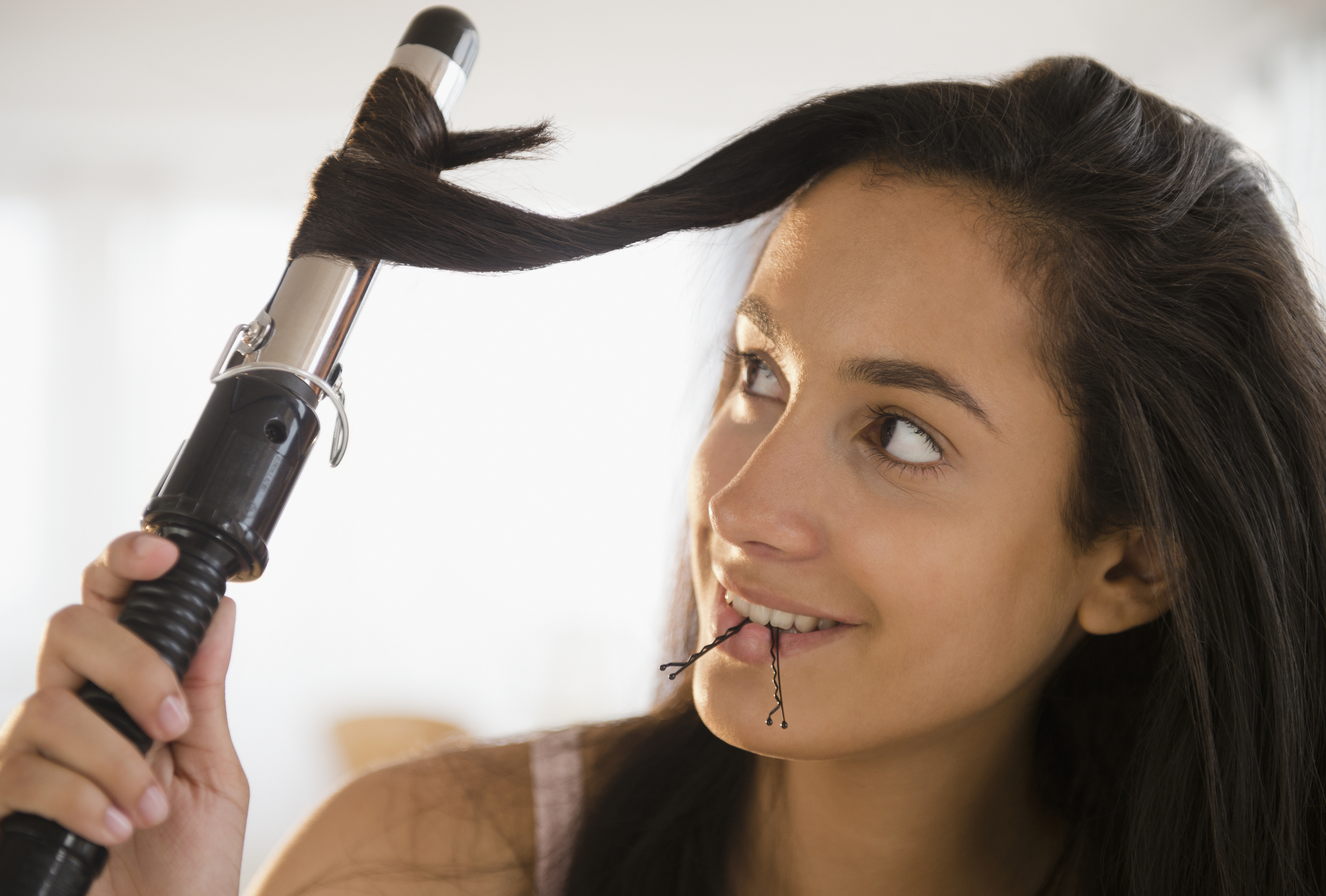 A person is using a curling iron on their hair while holding bobby pins in their mouth