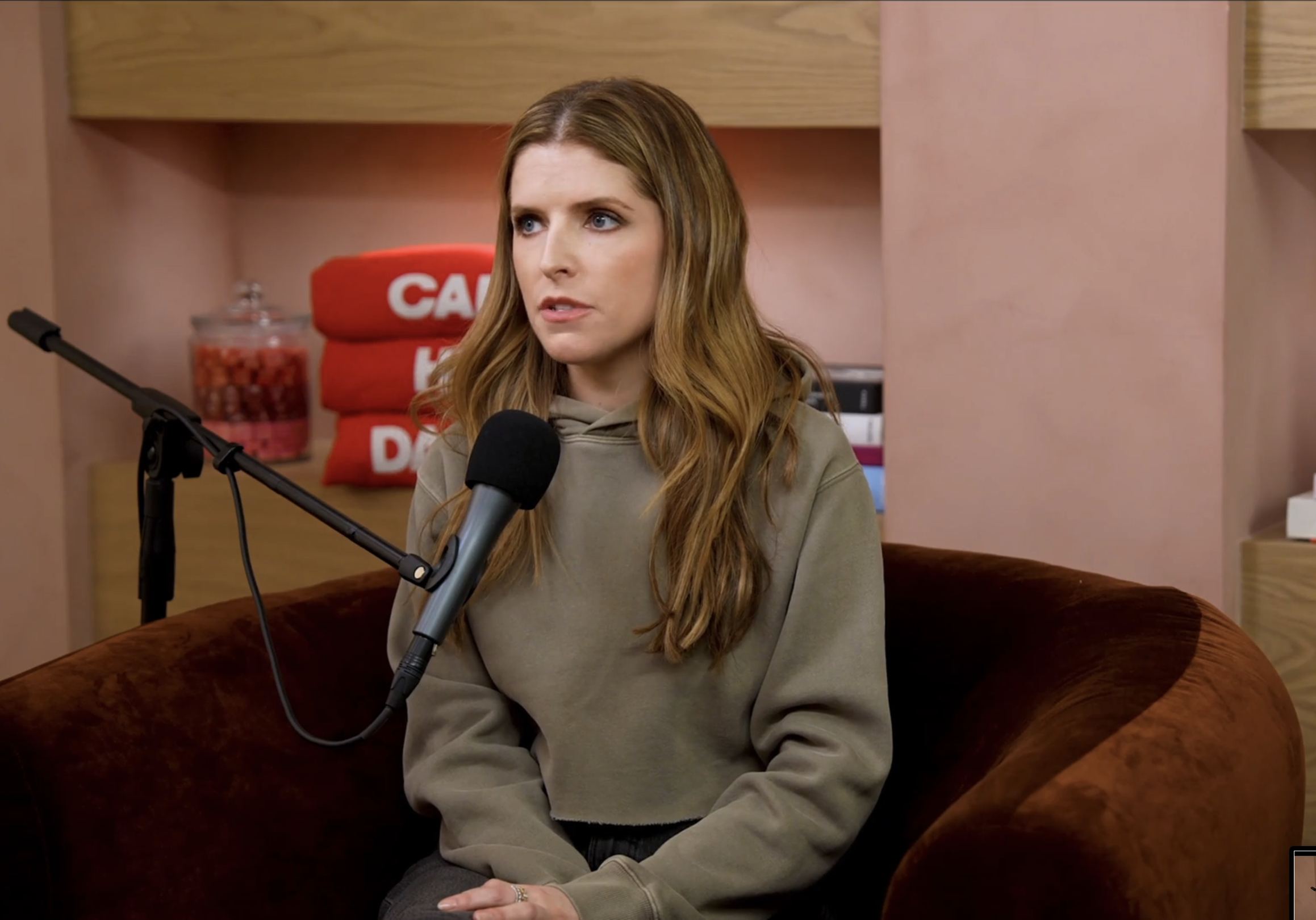 Anna in a cozy setting, sits in a chair, speaking into a microphone, with books and snacks visible on shelves behind her