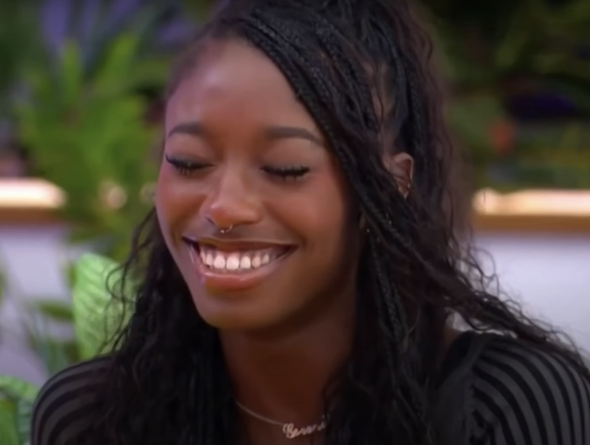 Smiling person with long braided hairstyle and septum piercing, wearing a striped top, with plants in the background