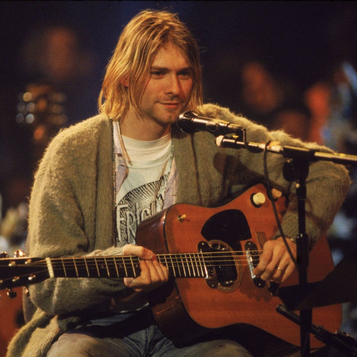 A person with long hair dressed in a t-shirt and cardigan plays an acoustic guitar on stage