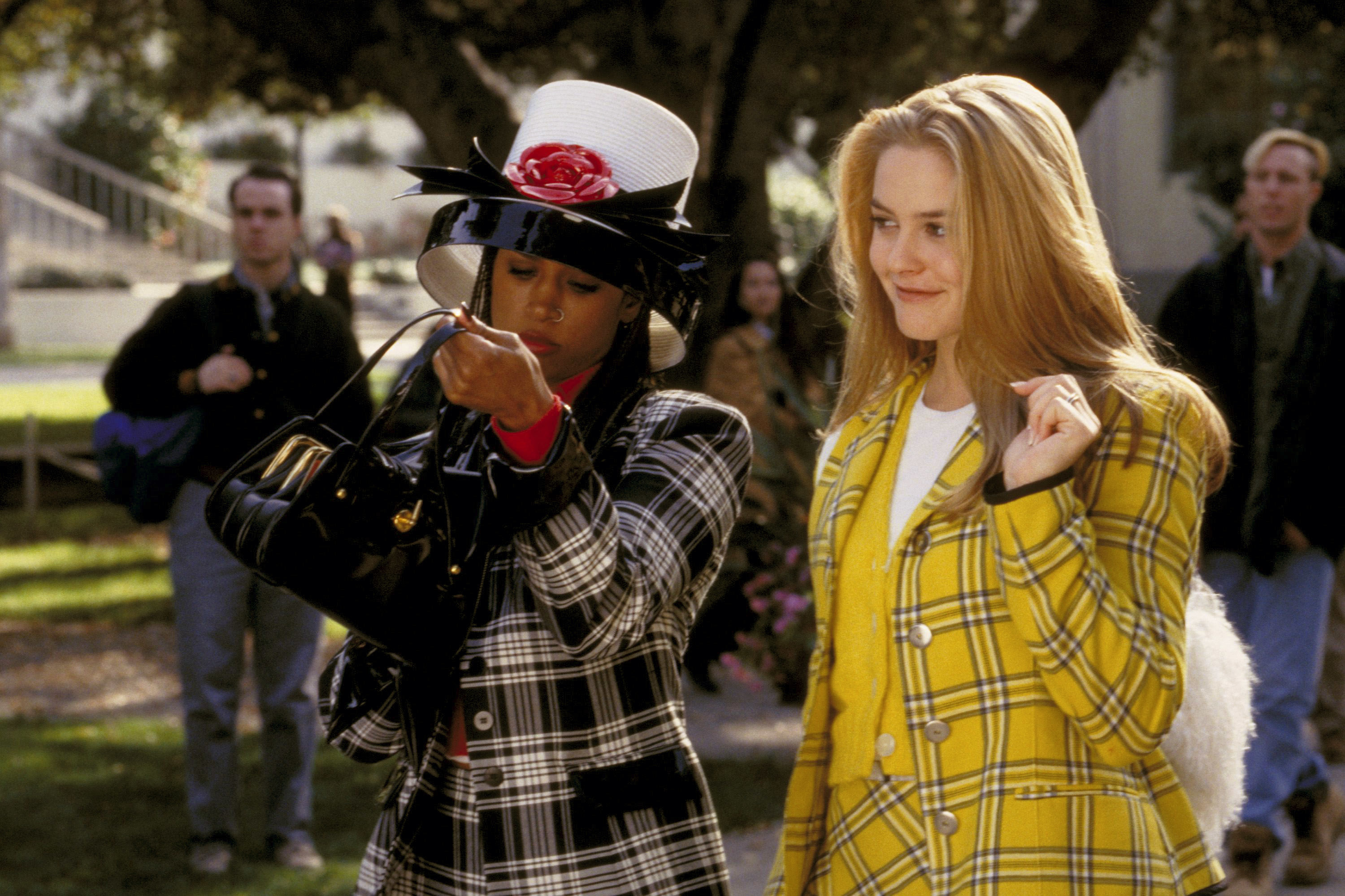 Two women wearing stylish plaid outfits; one with a white hat and rose, the other with a yellow jacket. Scene from "Clueless" movie